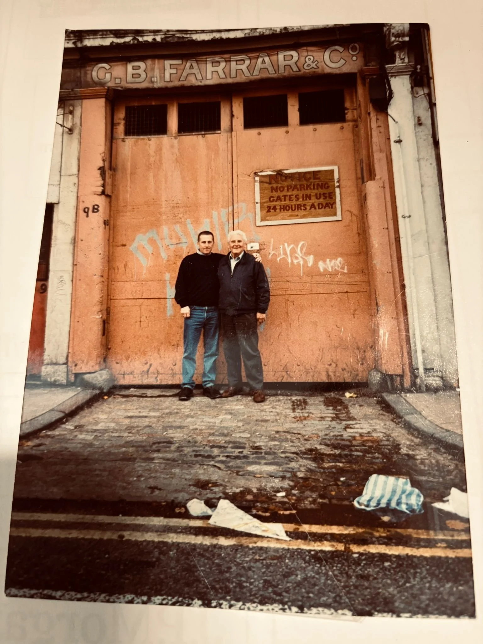 Two men standing in front of a large, closed, rusty metal door with graffiti and a sign that reads "No Parking GATES IN USE 24 HOURS A DAY." The ground is wet and cluttered with trash, including a crumpled piece of paper and a bag. One man has light hair and is wearing a dark jacket, while the other has dark hair and is wearing a black sweater.