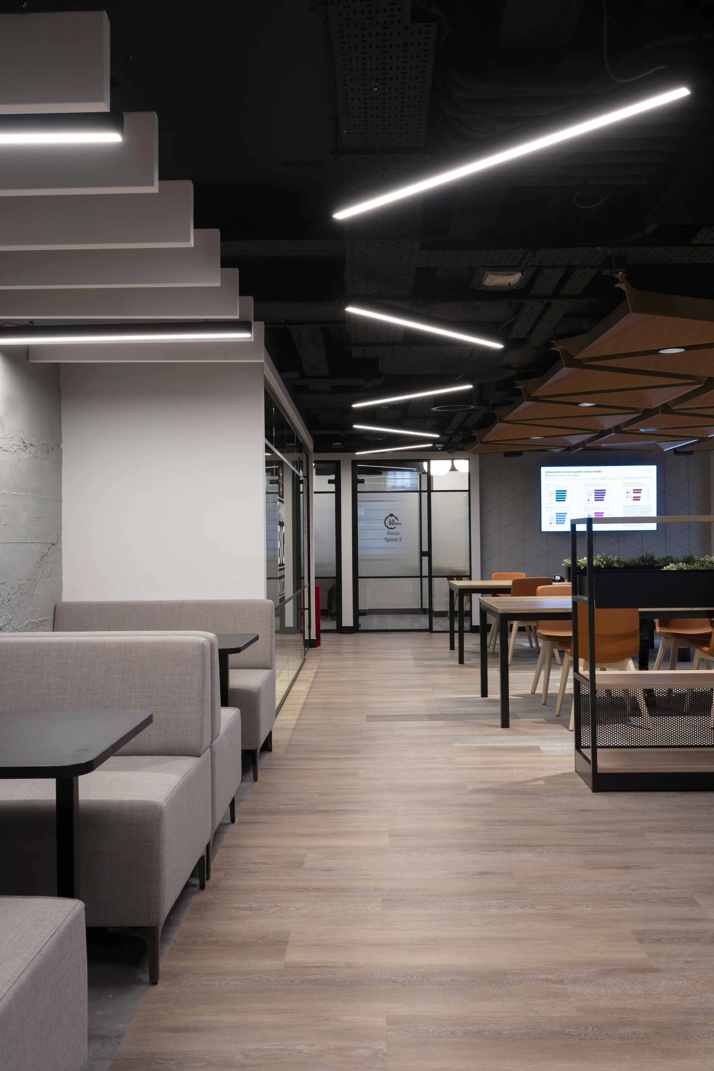Modern office lounge with gray sofas, wooden tables, dining chairs, a digital display on the wall, and contemporary ceiling lighting.