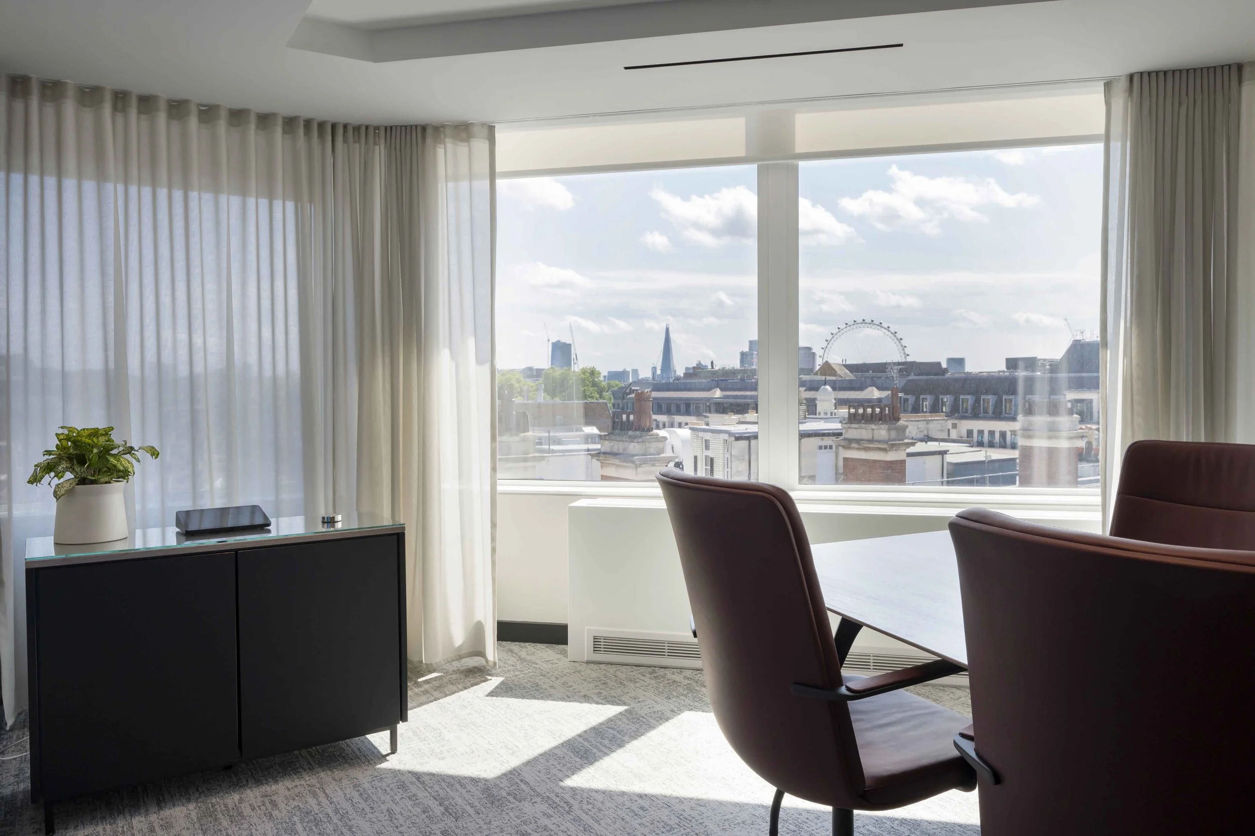 A modern office with large windows showing a city skyline including a Ferris wheel and skyscrapers, beige curtains, a black cabinet with a potted plant, and maroon chairs around a wooden table.