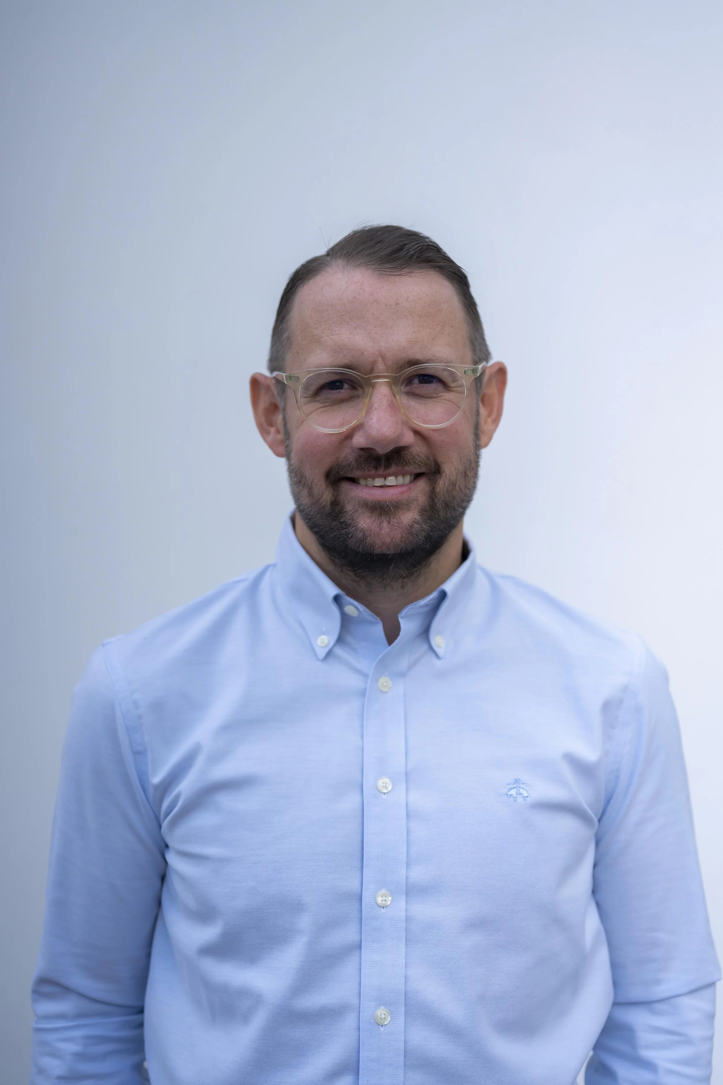 A man with short brown hair, beard, and glasses wearing a light blue button-up shirt and smiling, standing against a plain light gray background.