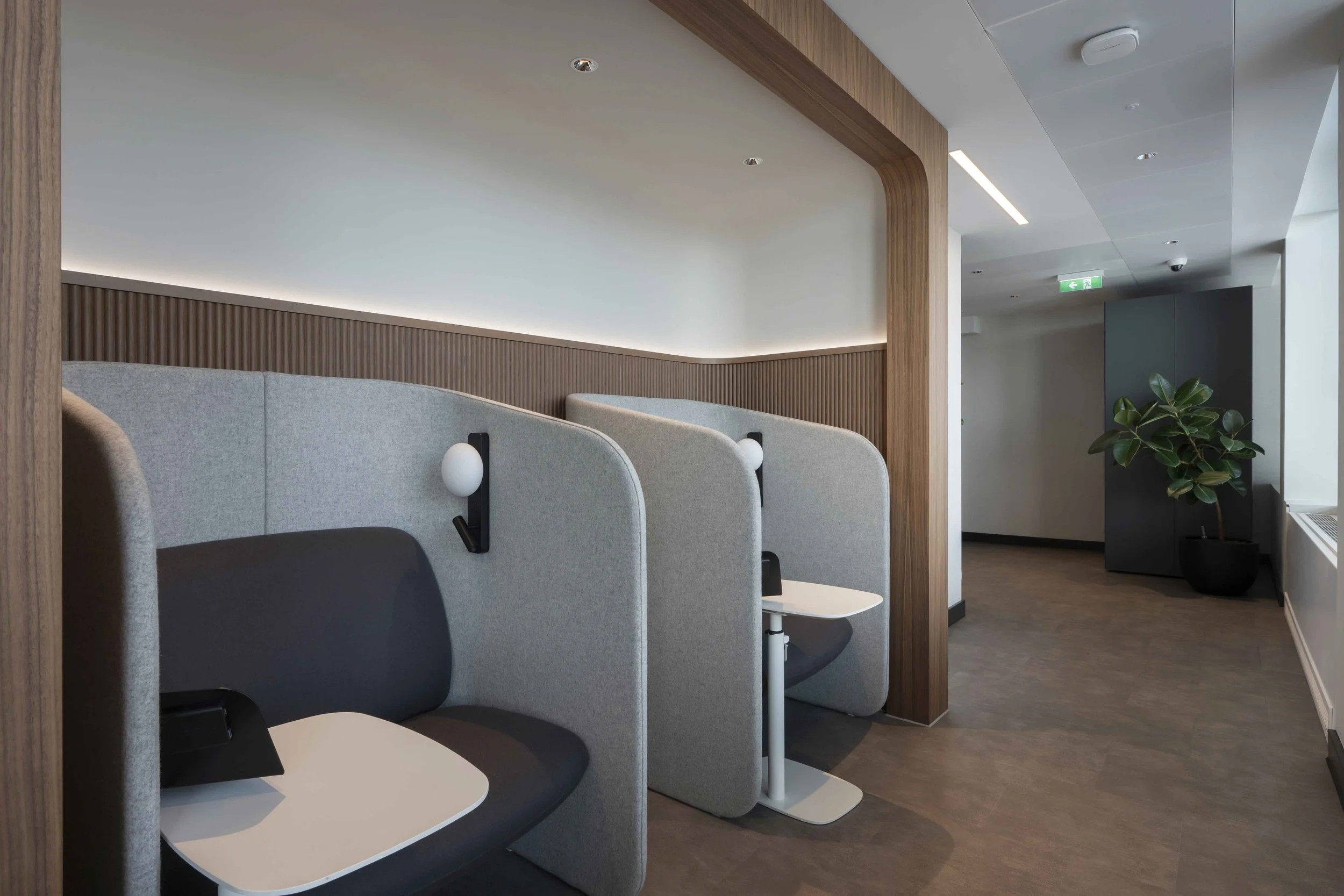 Interior of a modern waiting area with individual seats, wooden accents, a potted plant, and a hallway leading to an emergency exit.