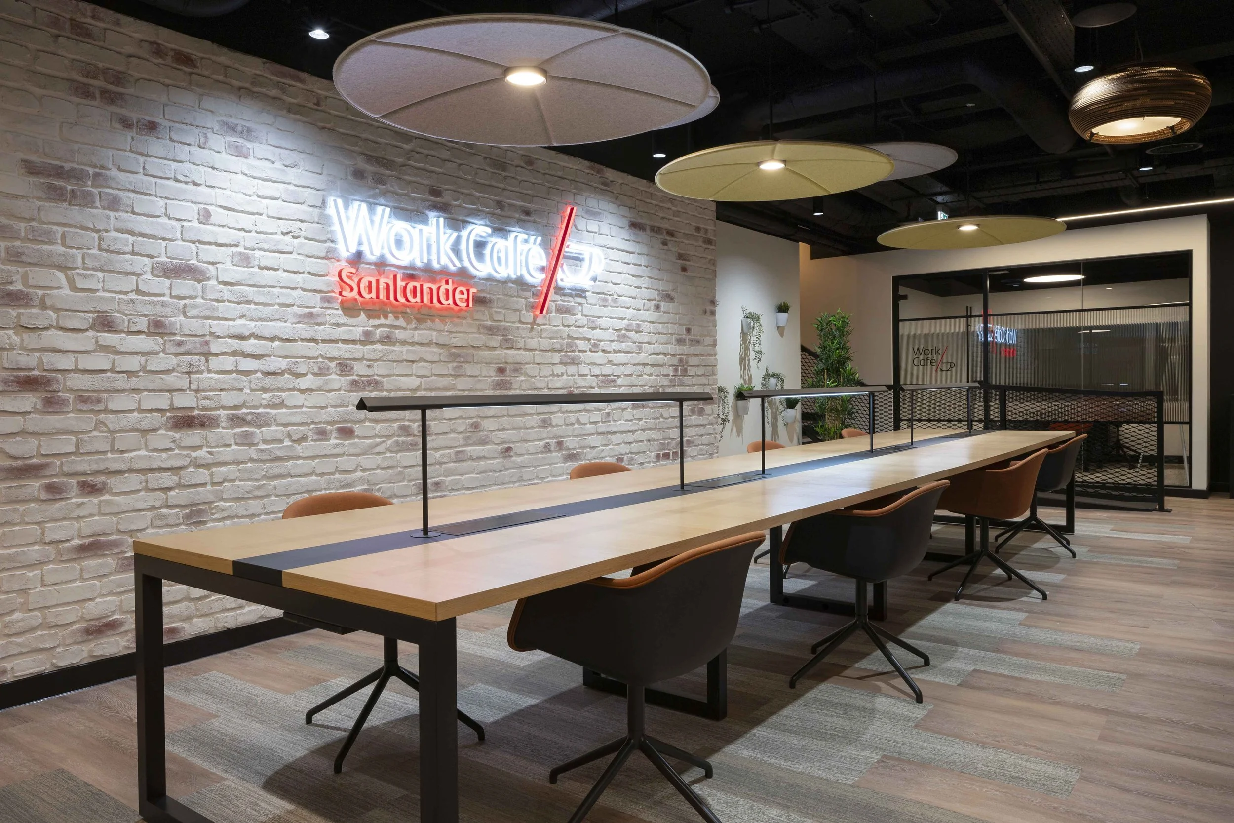 Interior of a modern office or coworking space with a long table, brown chairs, white brick wall, and a neon sign that says 'Work Cafe Santander'. Overhead lights and hanging decorative ceiling panels are also visible.
