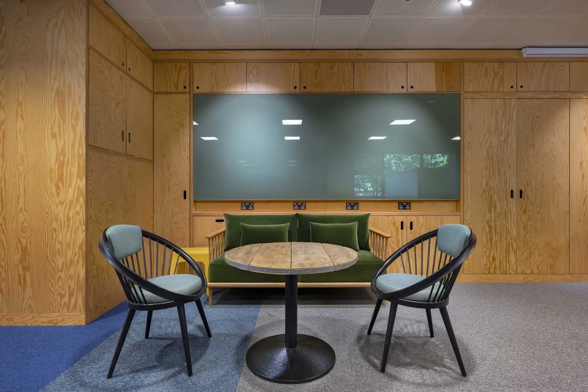 A meeting room with a round wooden table, four chairs with black frames and light blue cushions, a green couch with three green cushions, and wooden wall panels with cabinets. The room has a blue and gray carpet, a window reflecting trees, and electr
