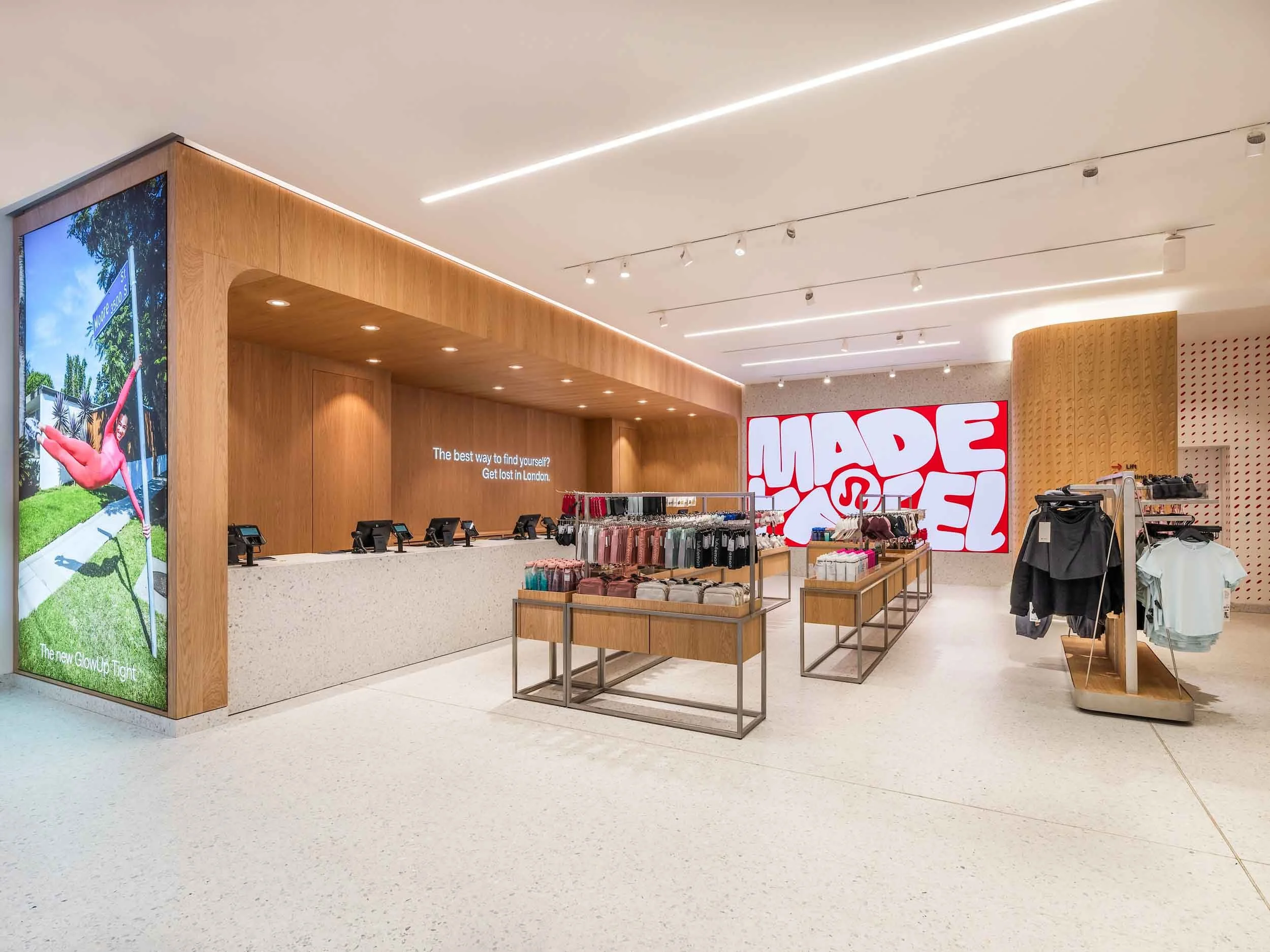 Interior of a retail store with wooden counters and displays of clothing, a large digital screen with a yoga pose, and a bright, modern ceiling with integrated lighting.