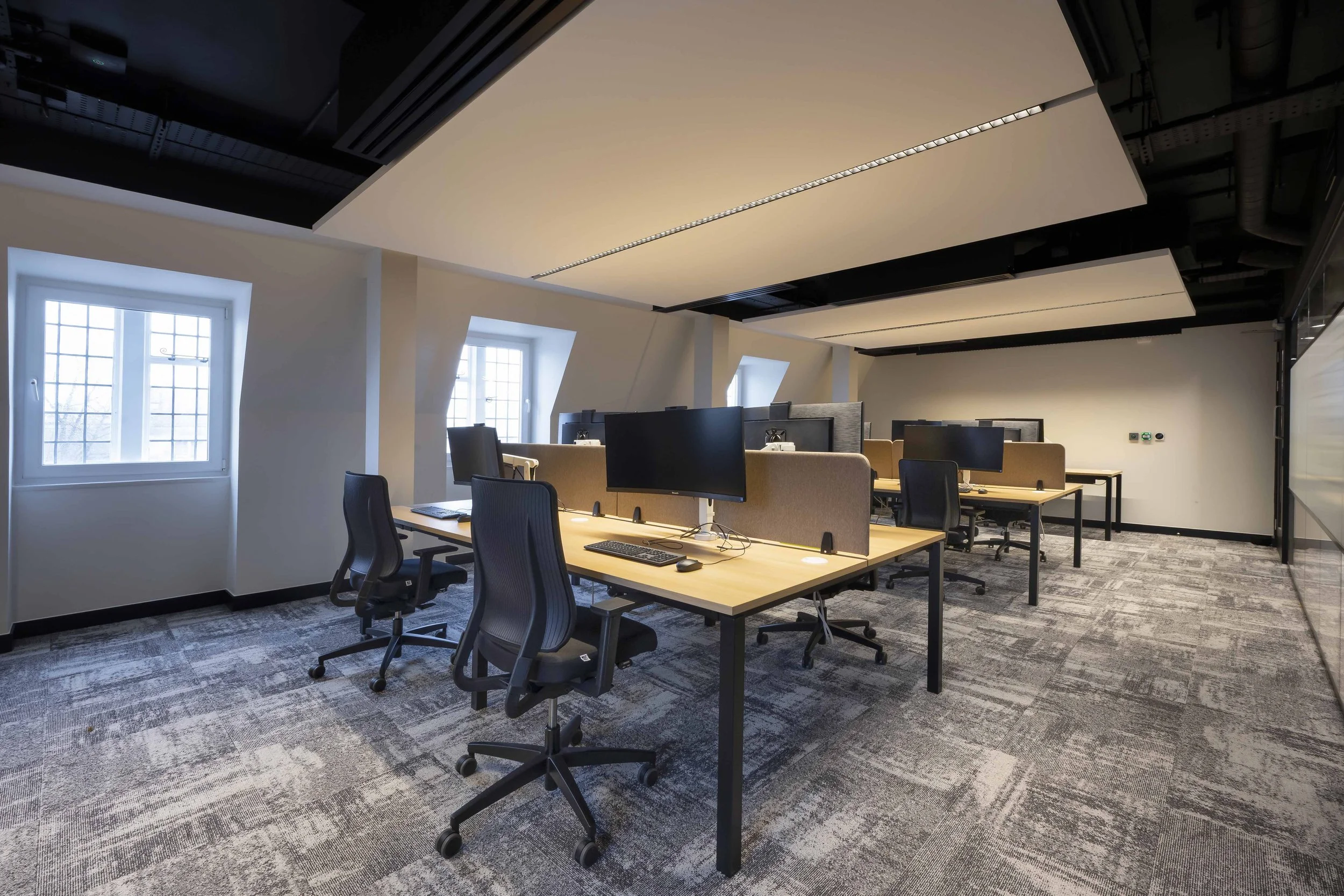Modern open-plan office with multiple desks, each equipped with a computer monitor, keyboard, and mouse. The room has large windows with grid-patterned panes allowing natural light, and a ceiling with black and beige panels and integrated lighting. The floor is covered with patterned gray carpet.