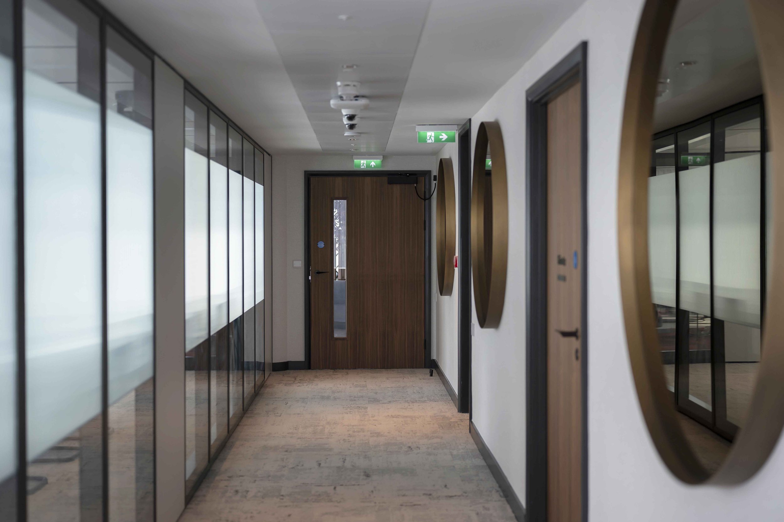 Modern office corridor with glass walls, wooden door at the end, overhead emergency exit signs, and ceiling cameras.