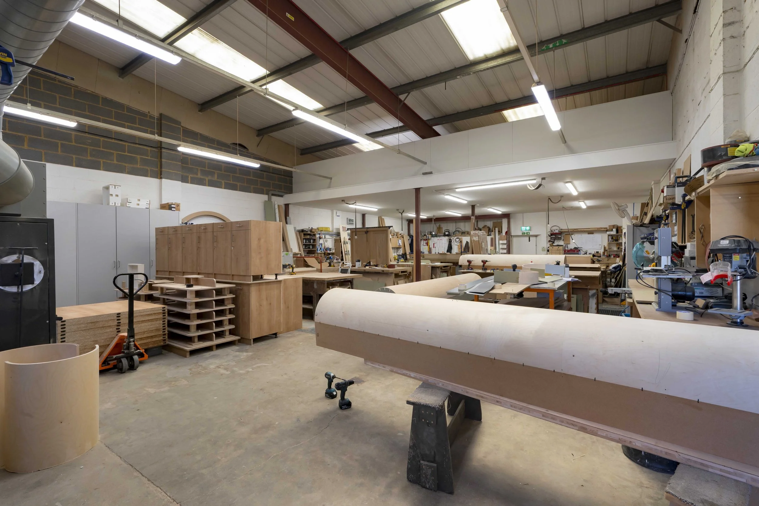 Interior of a woodworking workshop with various wooden furniture pieces, tools, and workbenches under a high ceiling with fluorescent lighting.