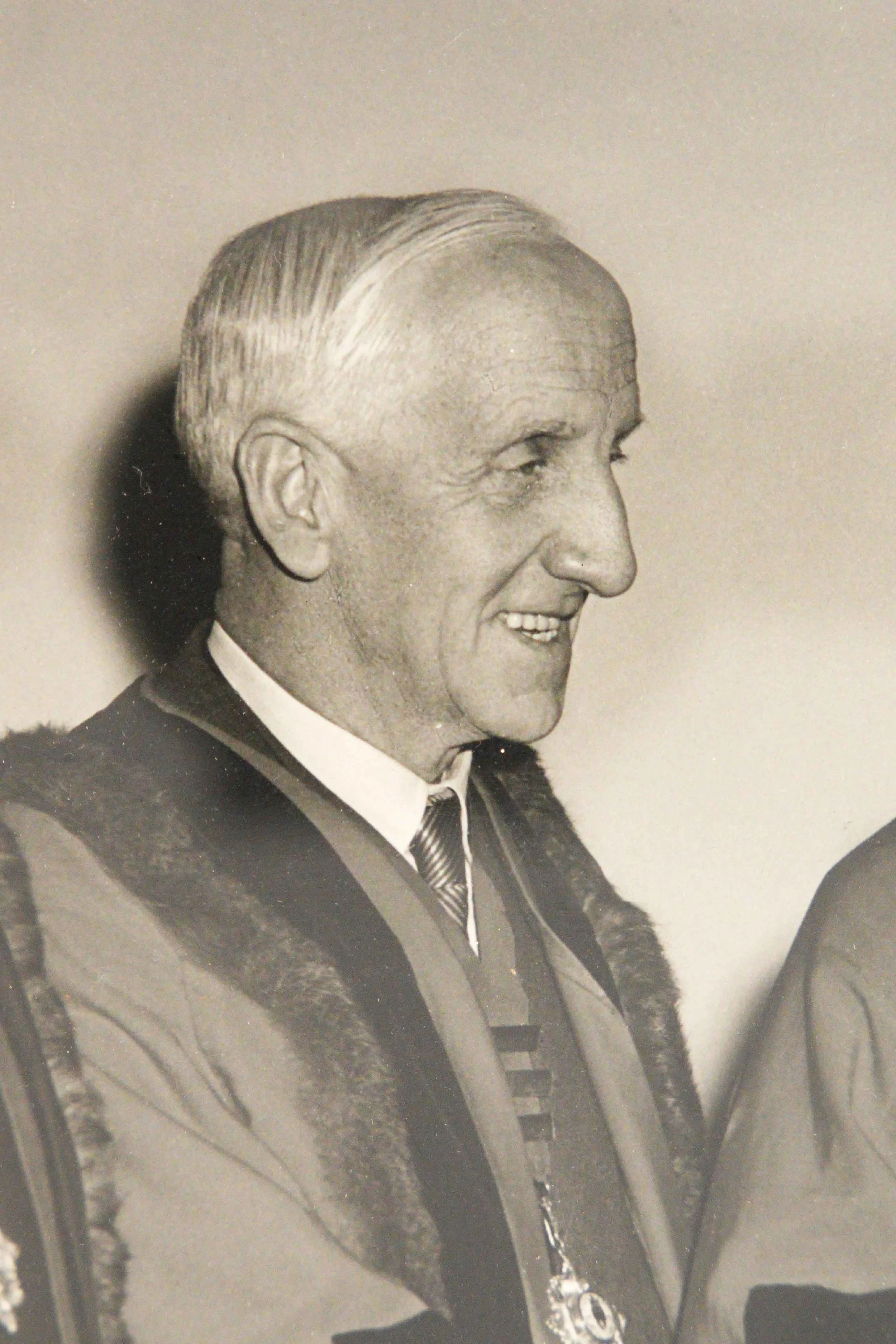 A black and white photograph of an older man with neatly combed back hair, smiling, dressed in a suit with a tie, and wearing a fur-collared coat.