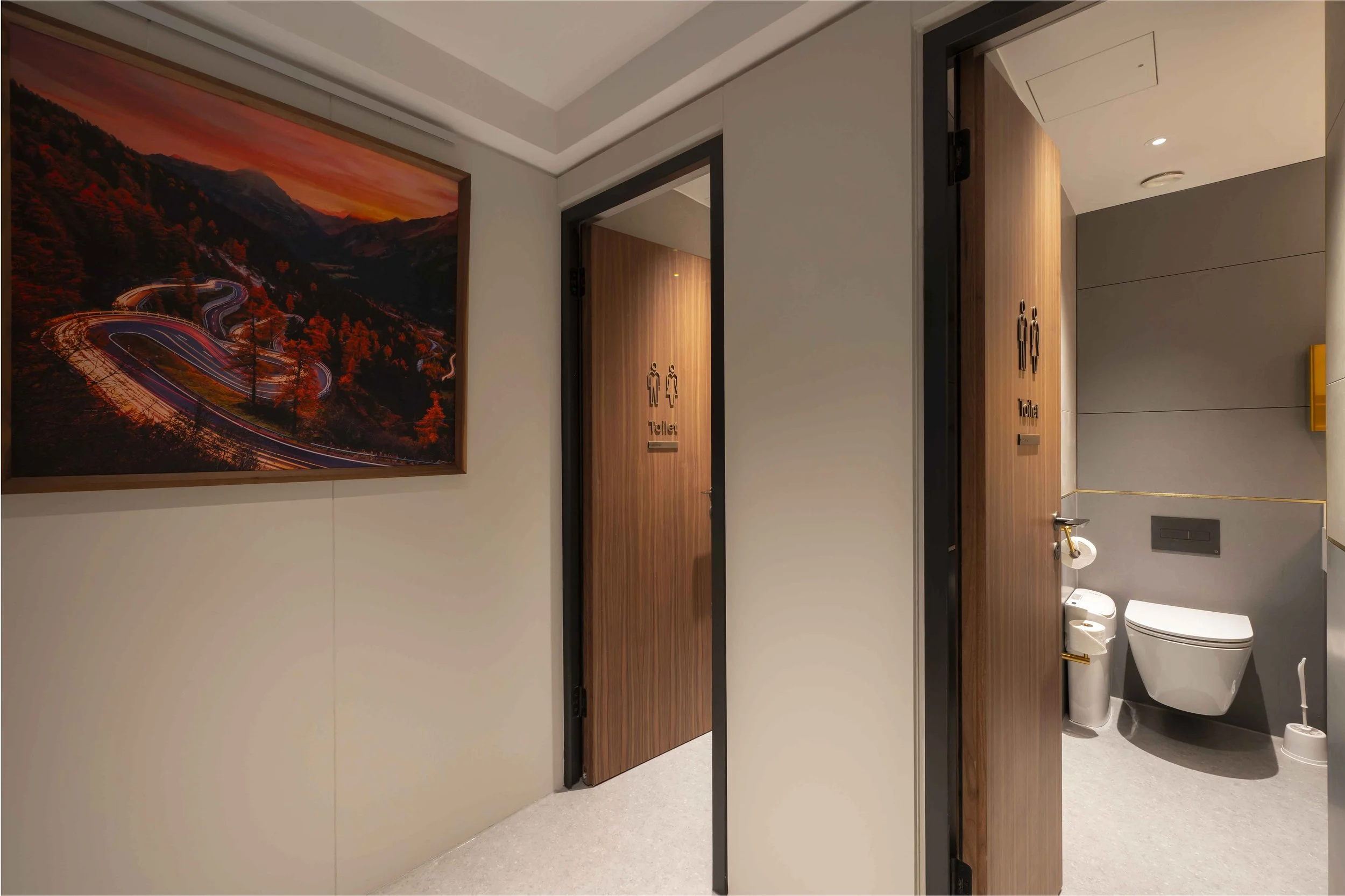 Public restroom with two doors labeled men's and women's toilets. Part of a wall with a large sunset mountain landscape painting is visible.