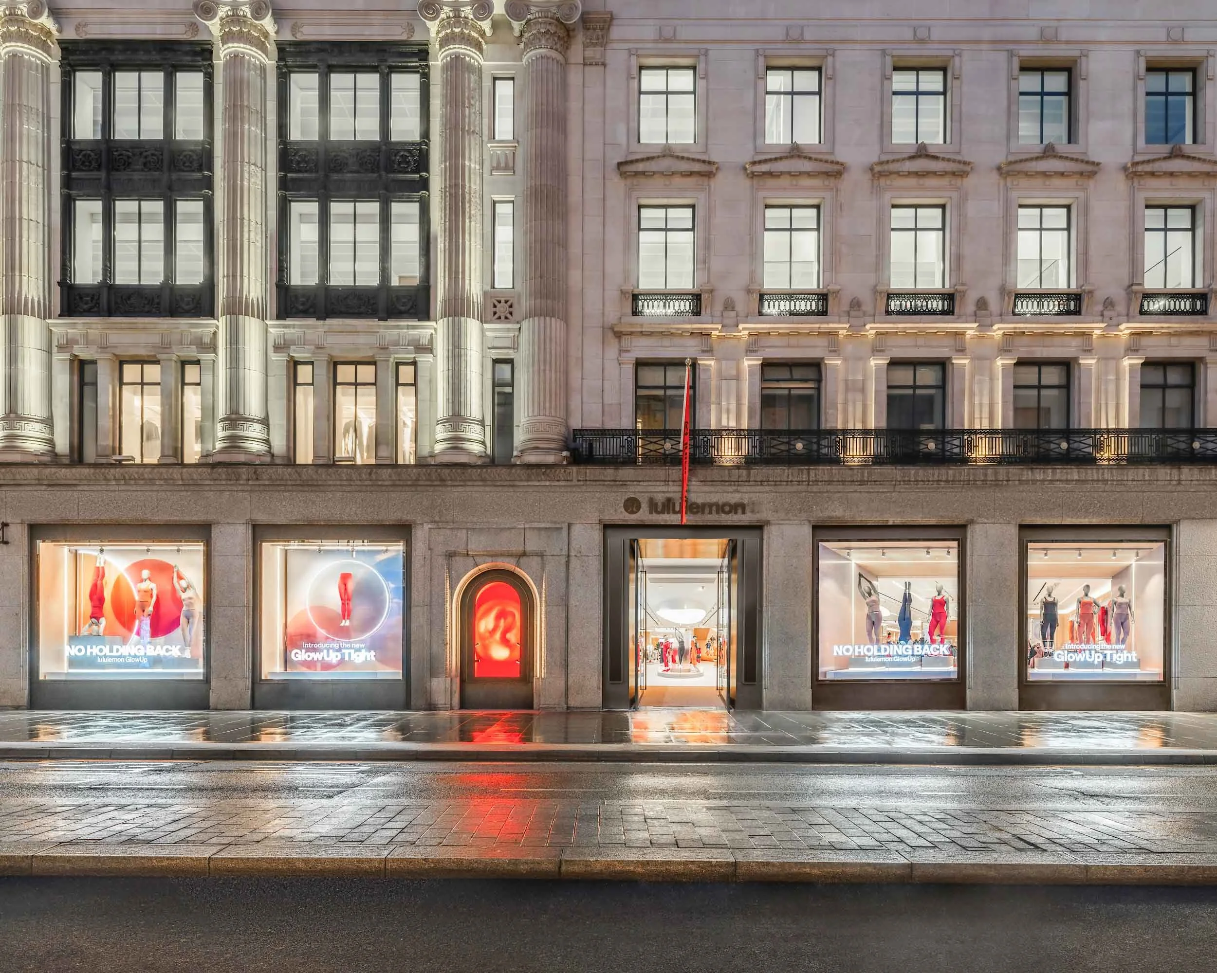 A multi-story building with retail stores on the ground floor, displaying mannequins dressed in clothing in the windows, illuminated at night. The building's facade has ornate columns and balconies, with a brick pattern on the upper levels.
