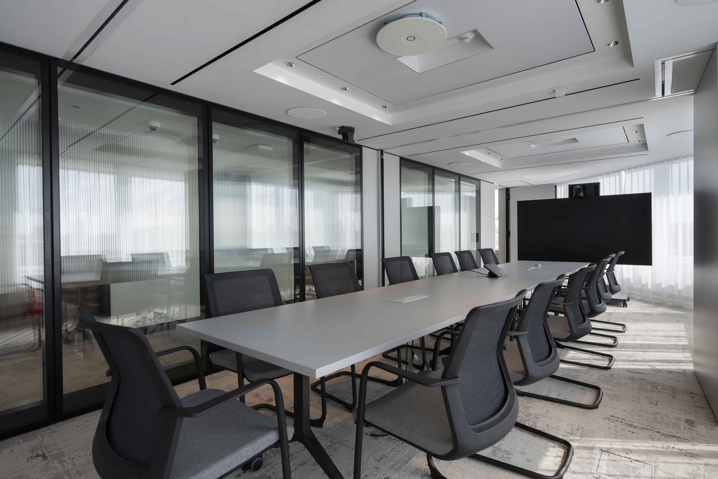 A modern conference room with a long white table, black mesh chairs, glass walls, and a large flat screen monitor at the end of the room.
