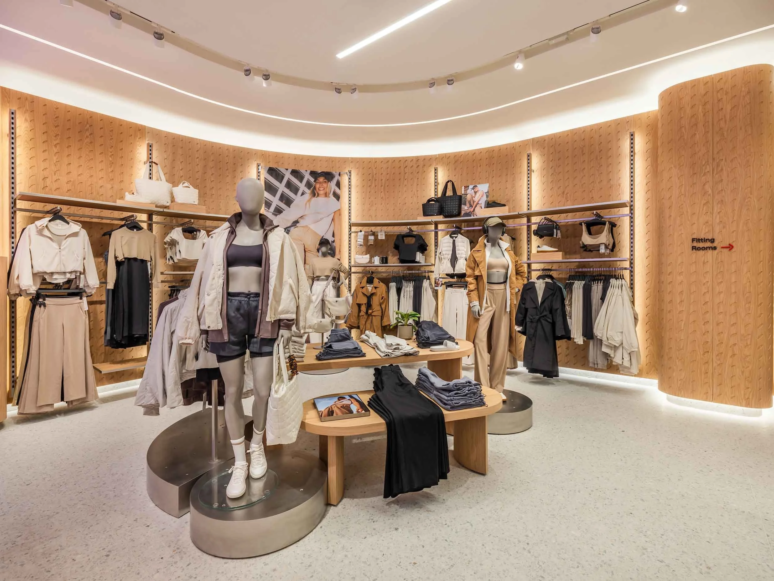 Clothing store display with mannequins dressed in casual and athletic wear, shelves with bags, books, and folded clothing, and promotional posters in the background.