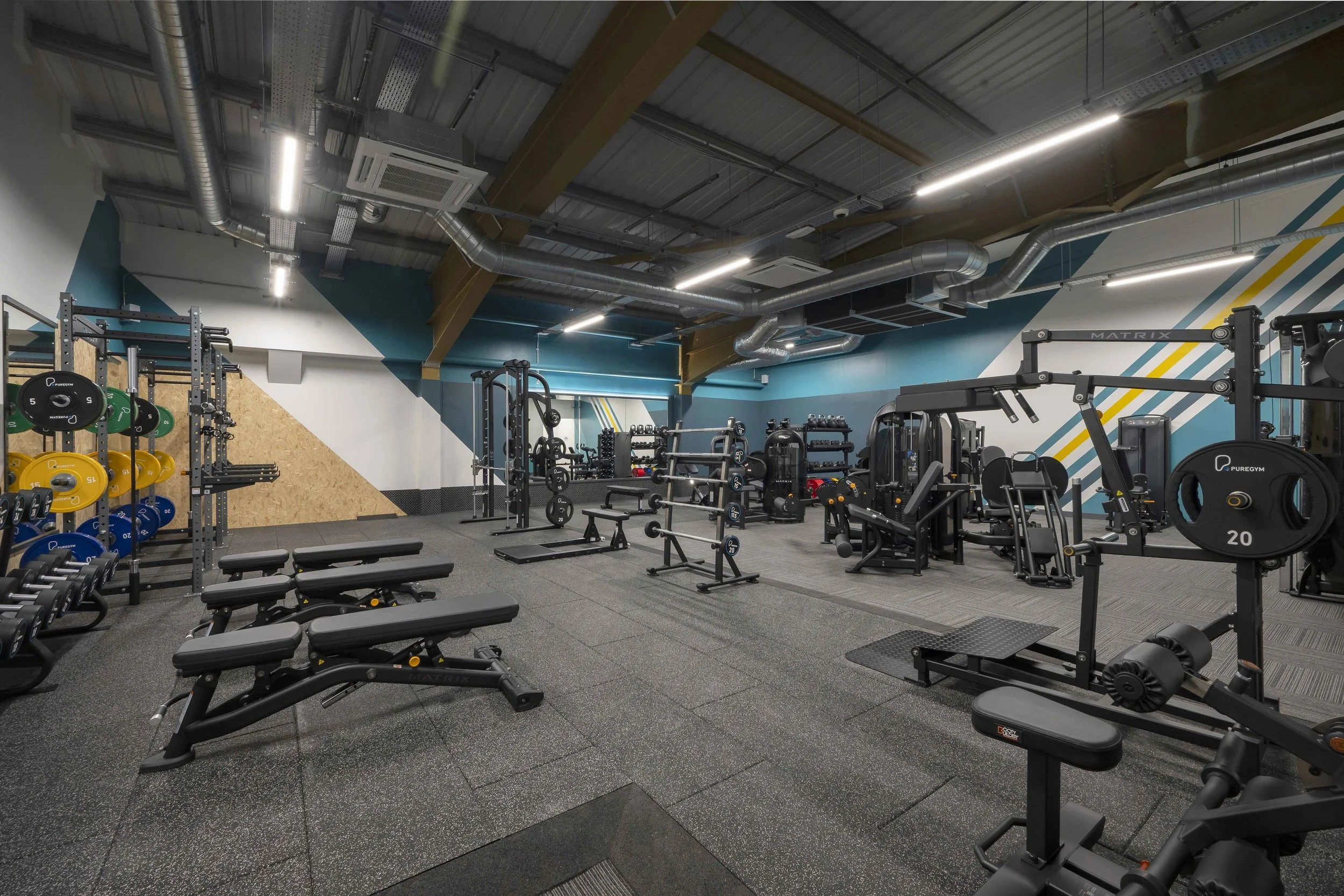 A modern gym with various workout equipment including dumbbells, benches, and weight machines, with a high ceiling and industrial-style exposed ductwork.