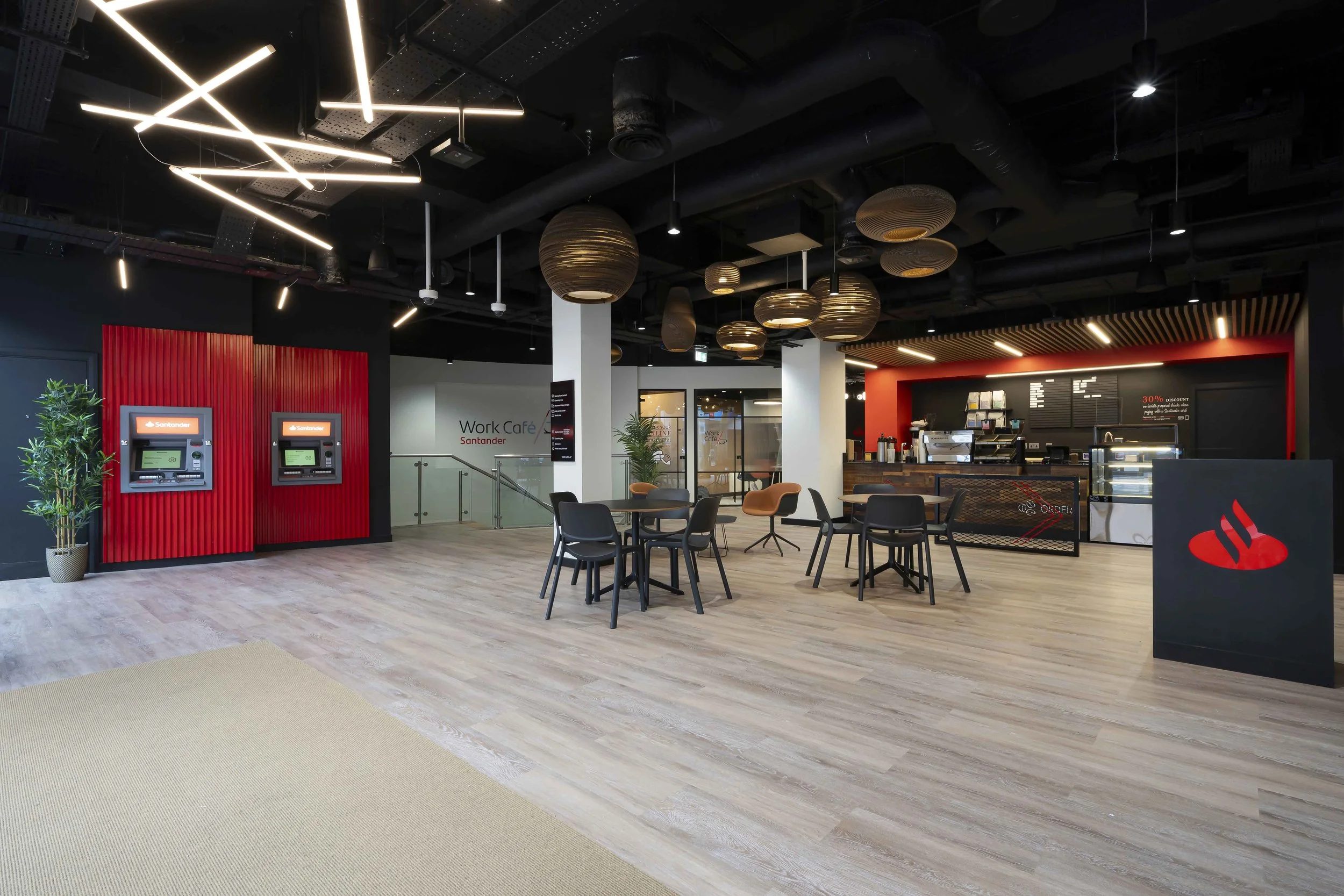 Interior of a modern coffee shop with black ceiling, artistic lighting fixtures, wooden accents, black tables and chairs, and a red wall with two ATMs. There is a counter area for ordering with menu boards and a display case.