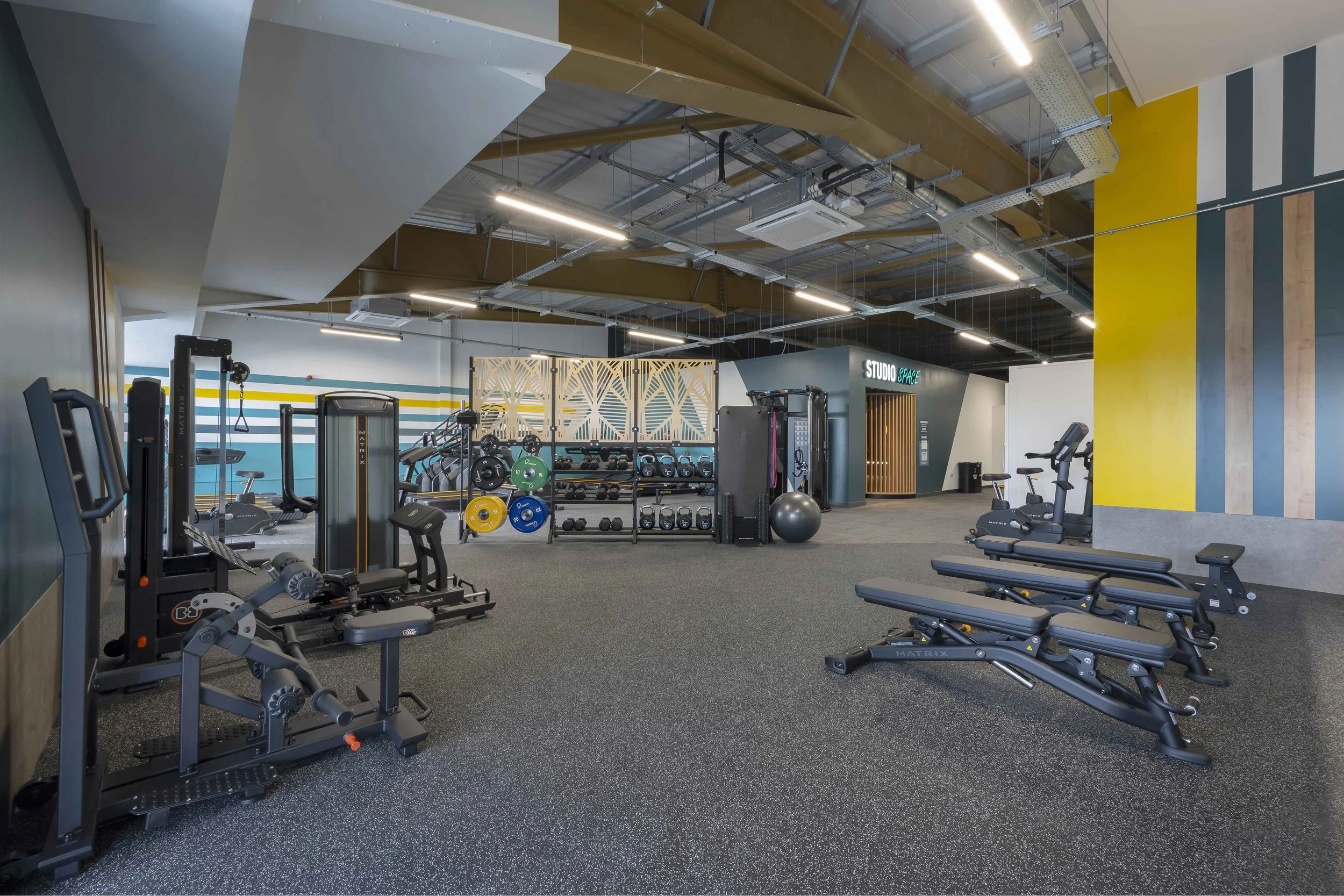Interior of a modern gym with workout equipment, including benches, weights, and cardio machines, with a colorful painted wall and an open ceiling featuring industrial-style piping and lighting.
