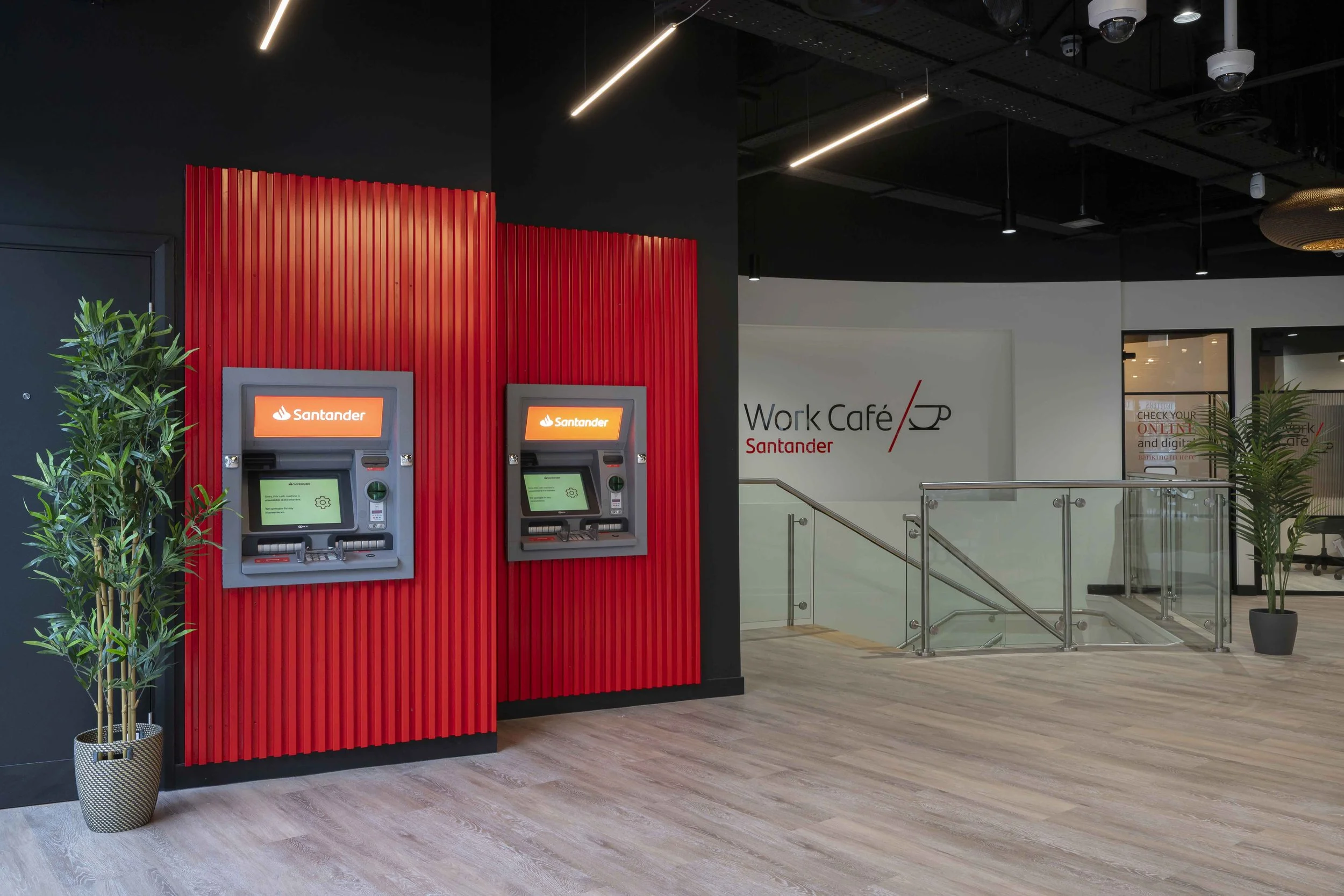 Indoor area with two ATMs on a red accent wall, green plants in pots, a white wall with 'Work Café Santander' signage, and a staircase leading up to an office space.