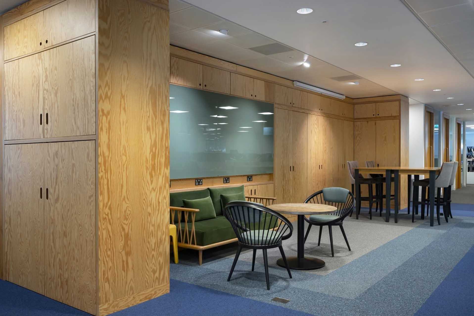 Modern office lounge area with wooden cabinets, a green sofa, round tables, and chairs in an open-plan space.