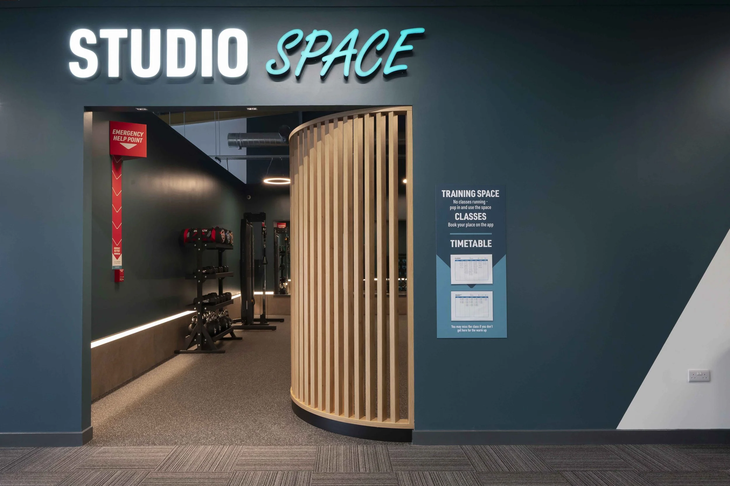 Entrance to a fitness studio named 'Studio Space' with a wooden folding gate, gym equipment inside, and signs indicating training space, classes, and timetable.