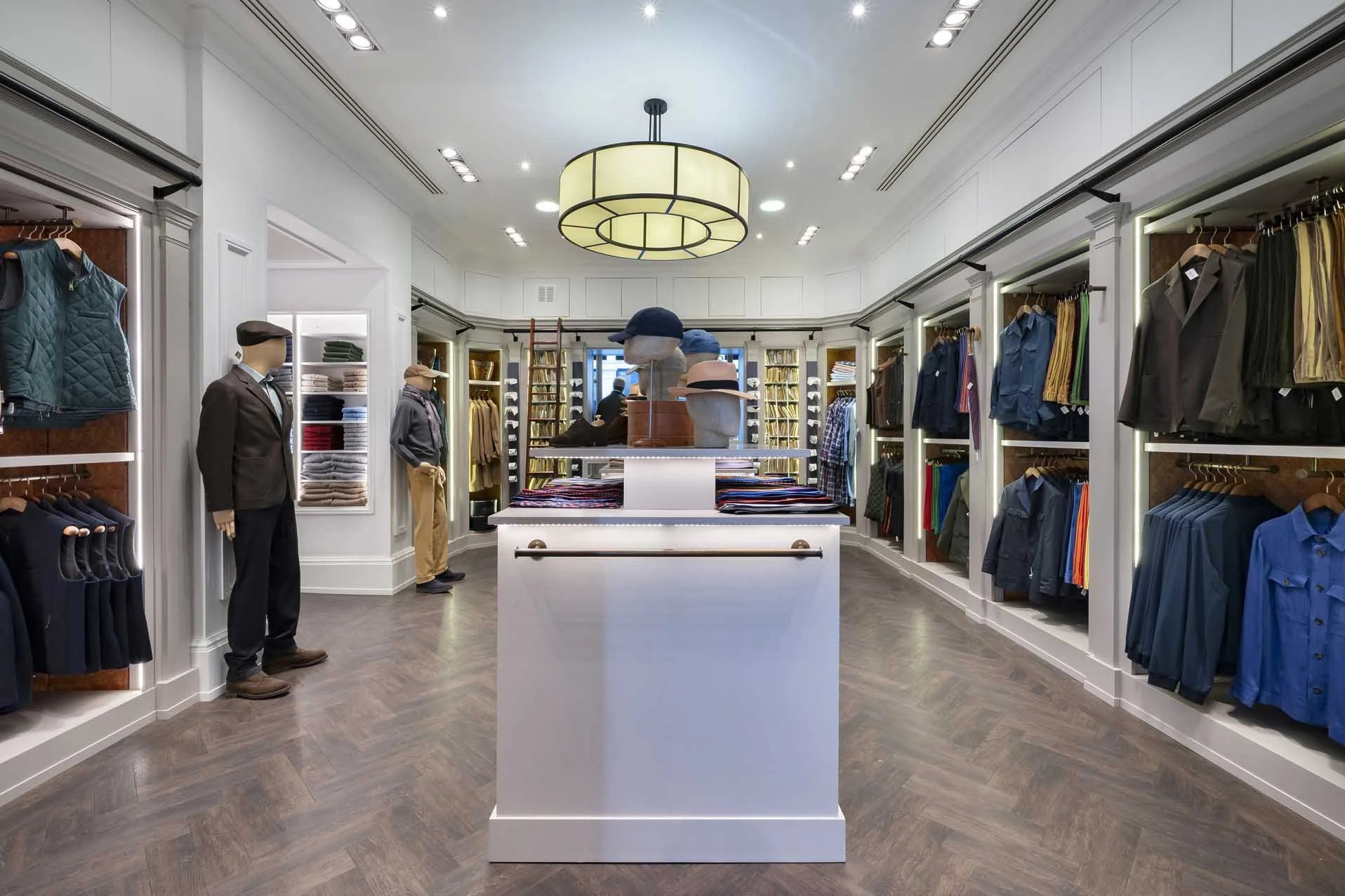 Interior of a clothing store with mannequins, shelves of folded clothes, hanging shirts and pants, and a central display table, lit by ceiling lights and a large circular chandelier.