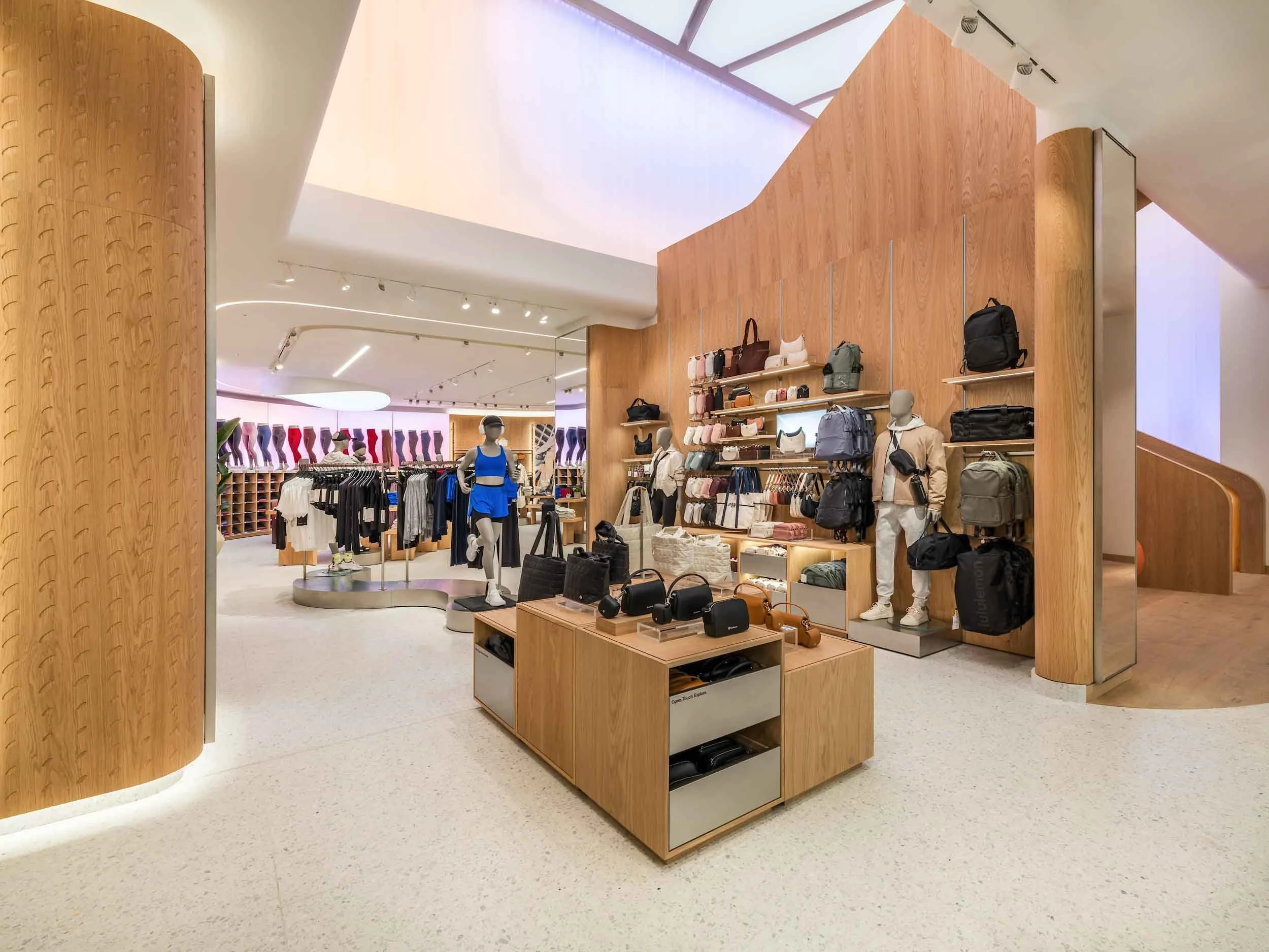 Interior of a modern retail store selling handbags, backpacks, and clothing, with mannequins and wooden display shelves, illuminated by natural and artificial light.
