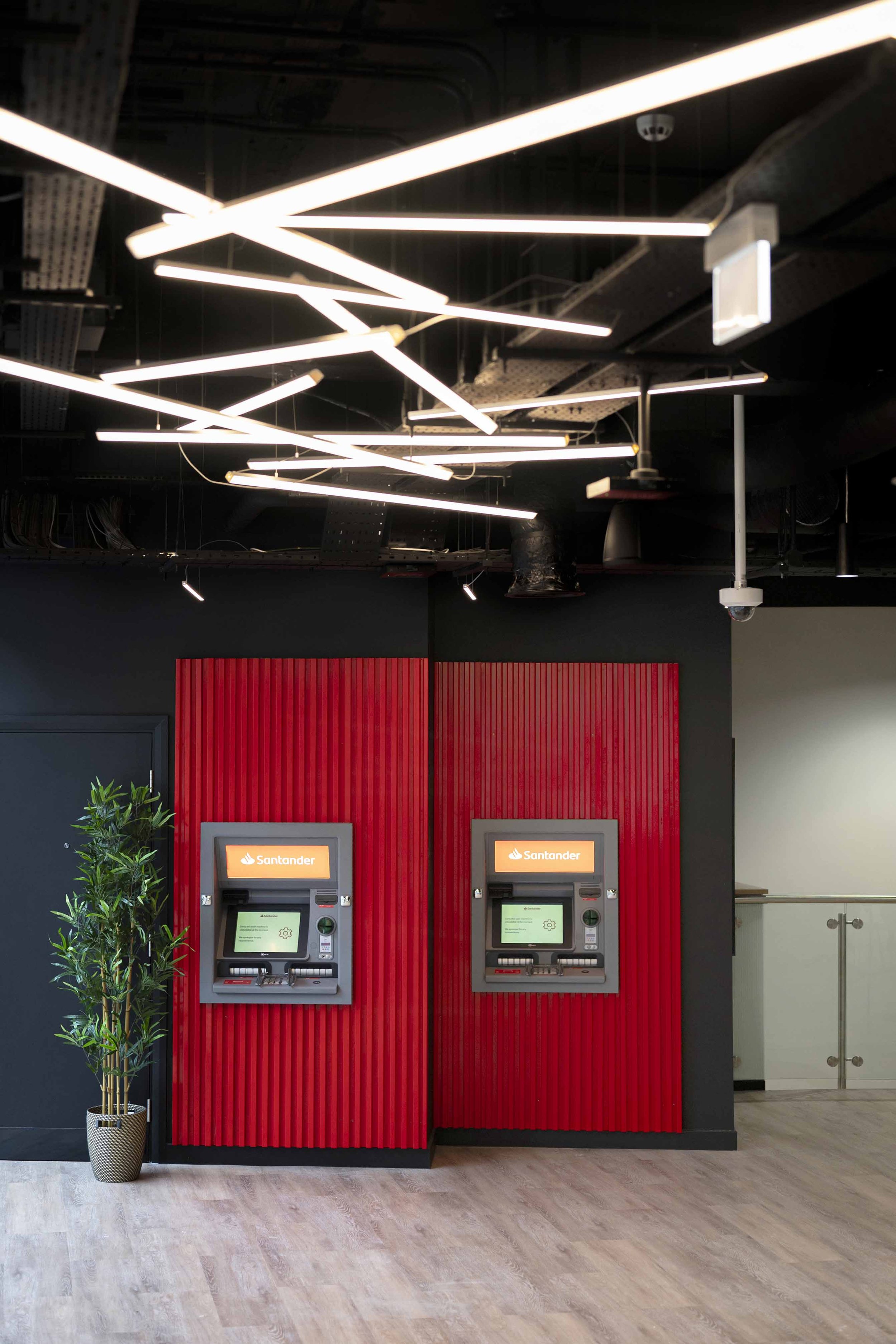 Two Santander ATMs against a red textured wall in a modern office or bank setting with wood flooring, a potted plant, and a black ceiling with geometric light fixtures.