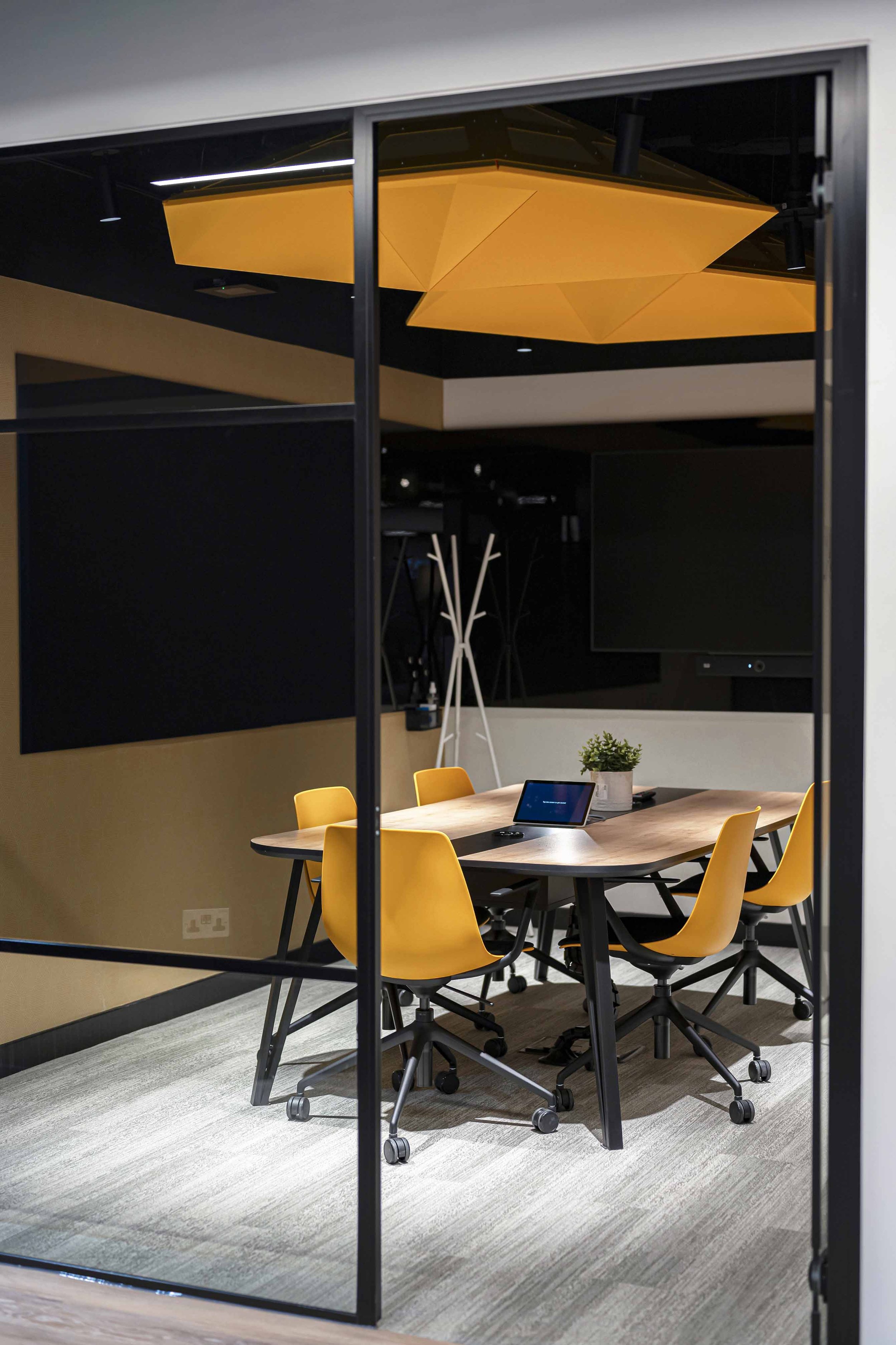 A conference room with a wooden table and yellow chairs, a potted plant, a tablet, a monitor, and a black wall with a large blank screen. The room is viewed through a glass wall with a black frame and a large yellow geometric ceiling light.