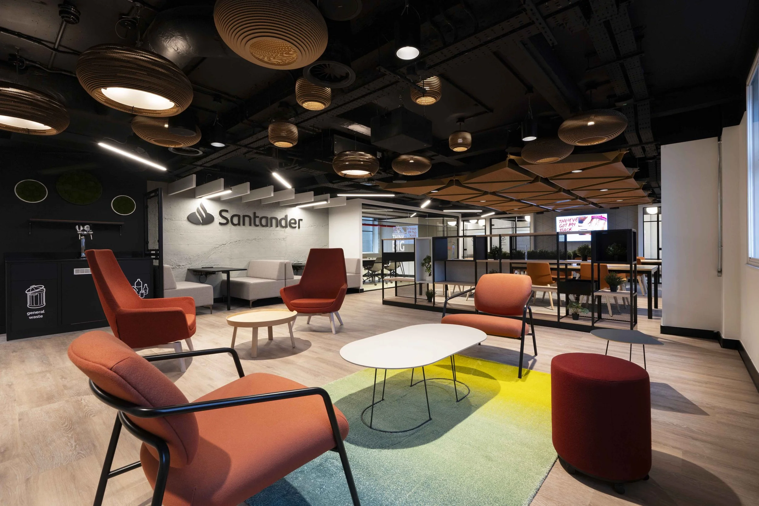 Modern office lounge area with orange and beige chairs, a small round table, a colorful rug, and a Santander sign on the wall. Ceiling features decorative lighting fixtures and wood paneling.