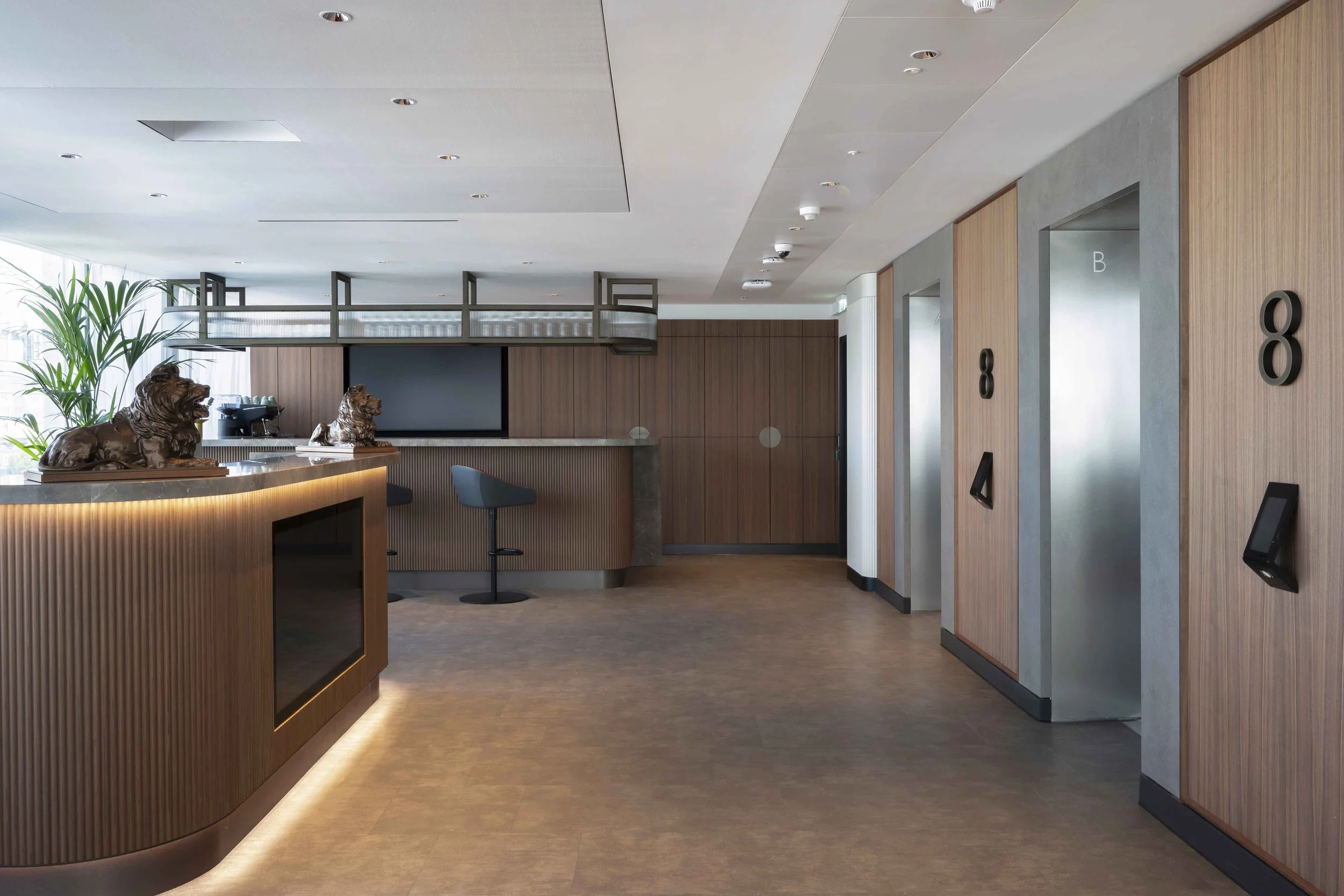 Modern hotel lobby reception area with a curved wooden front desk, decorative lion sculptures, green plants, and digital check-in kiosks along the wooden wall with room numbers.