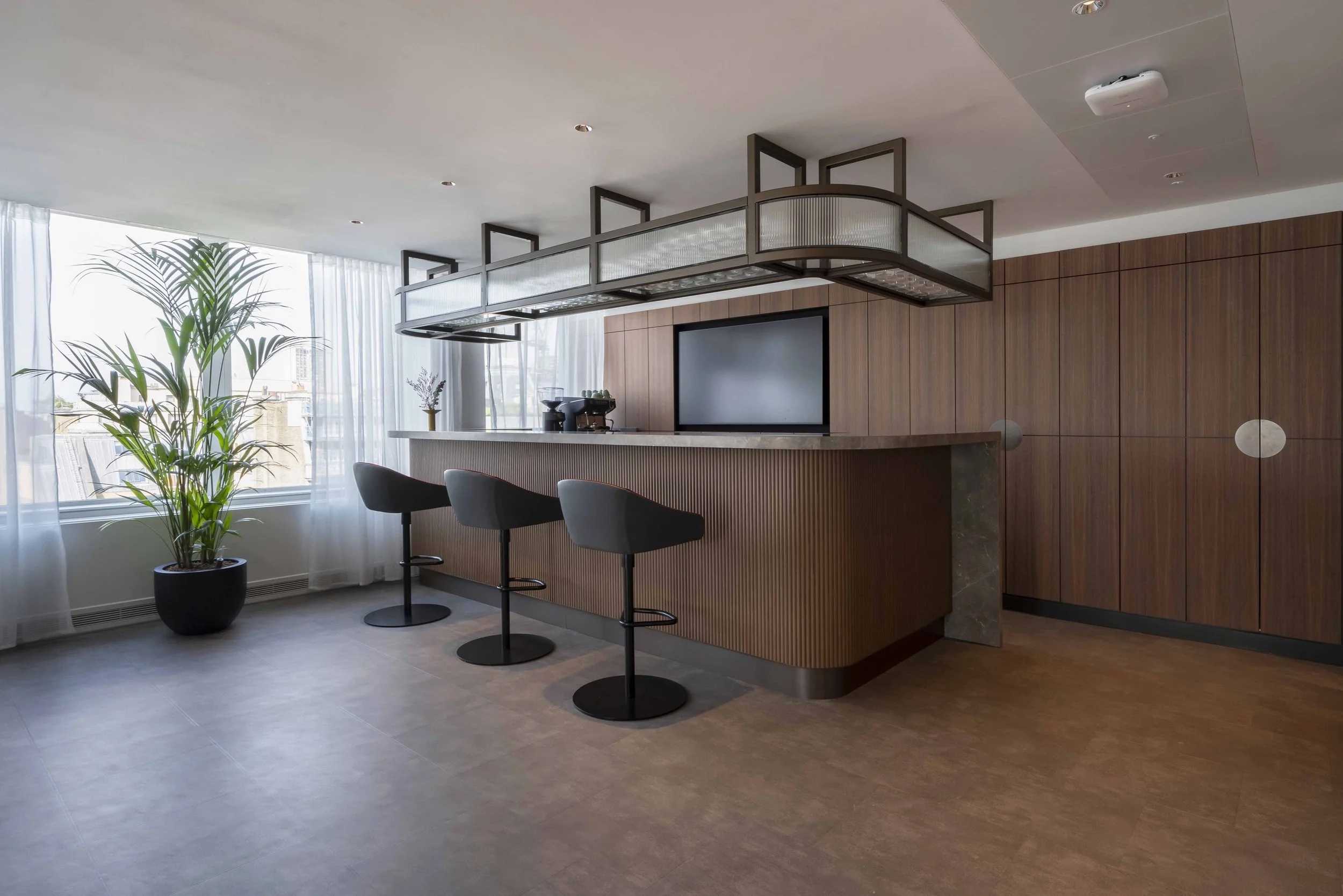 Modern hotel or apartment lounge area with a bar counter, three bar stools, a large window with sheer curtains, tall potted plant, and wood-paneled wall.