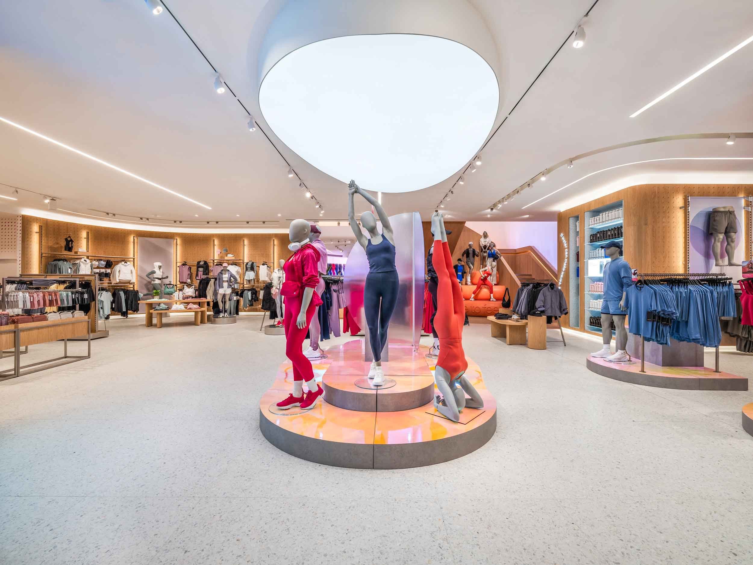 A clothing store display with mannequins dressed in athletic wear, surrounded by racks of clothes and wooden shelving in a modern retail space with curved ceiling design and bright lighting.