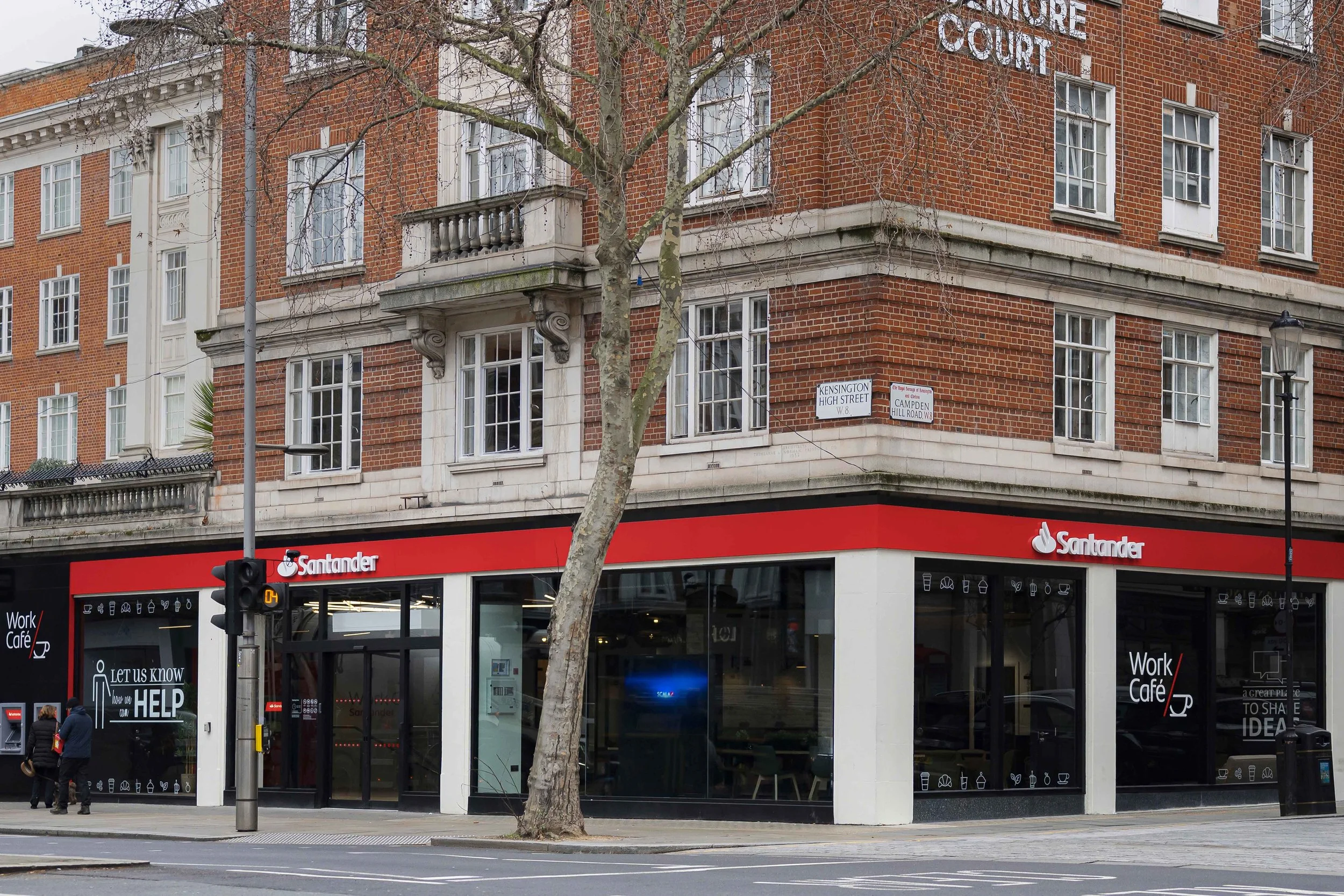 Street view of a brick building with a Santander bank branch on the ground floor, a tree in front, and pedestrians crossing the street near a traffic light.