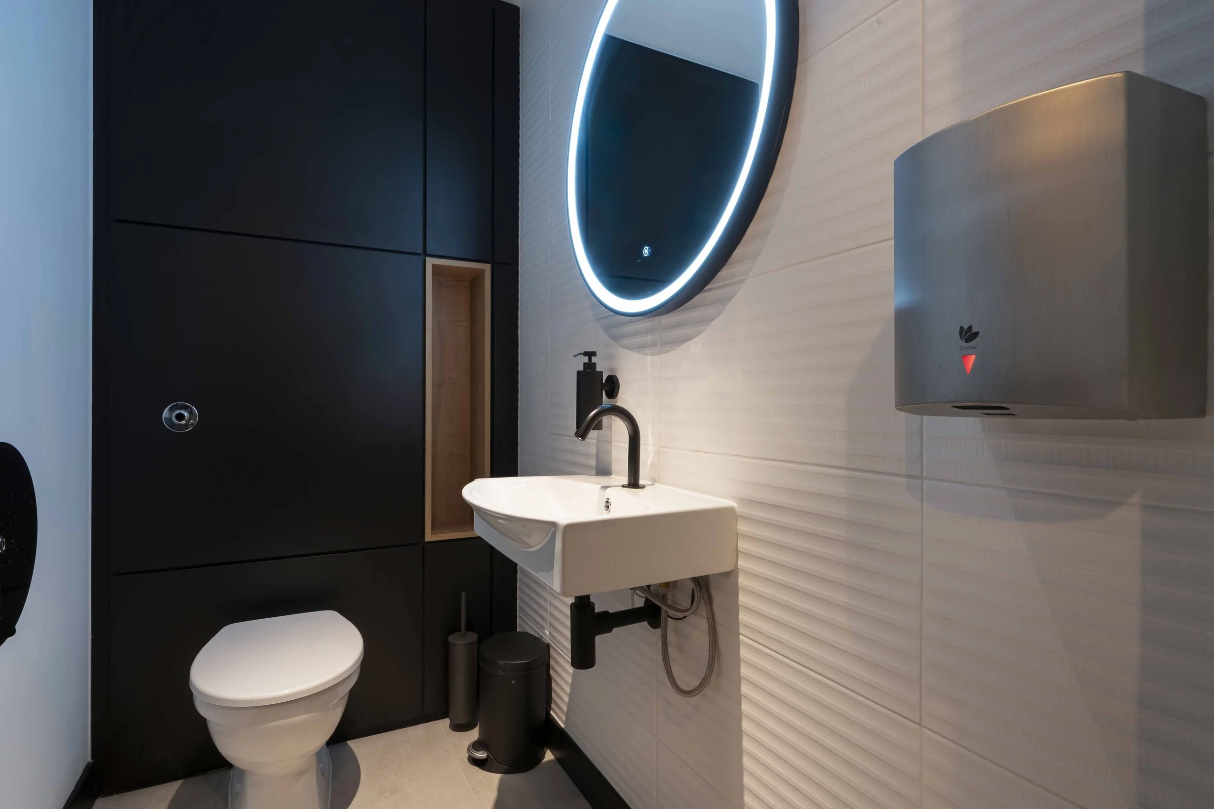 Modern bathroom featuring a white toilet, small sink with black fixtures, black cabinets, a unique illuminated oval mirror, and a hand dryer on the wall.
