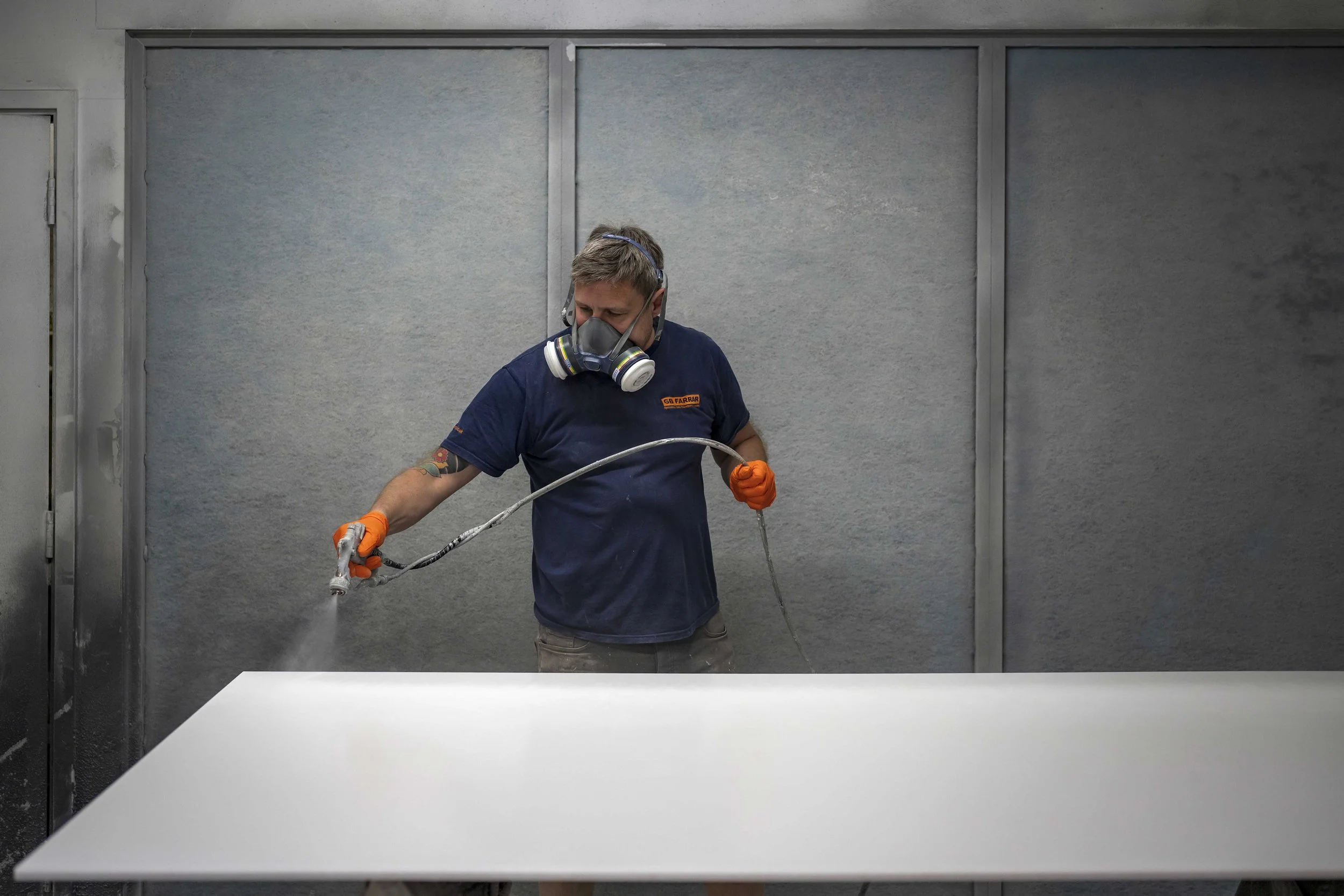 A man wearing a respirator mask and orange gloves is spray painting a white surface in an industrial setting.