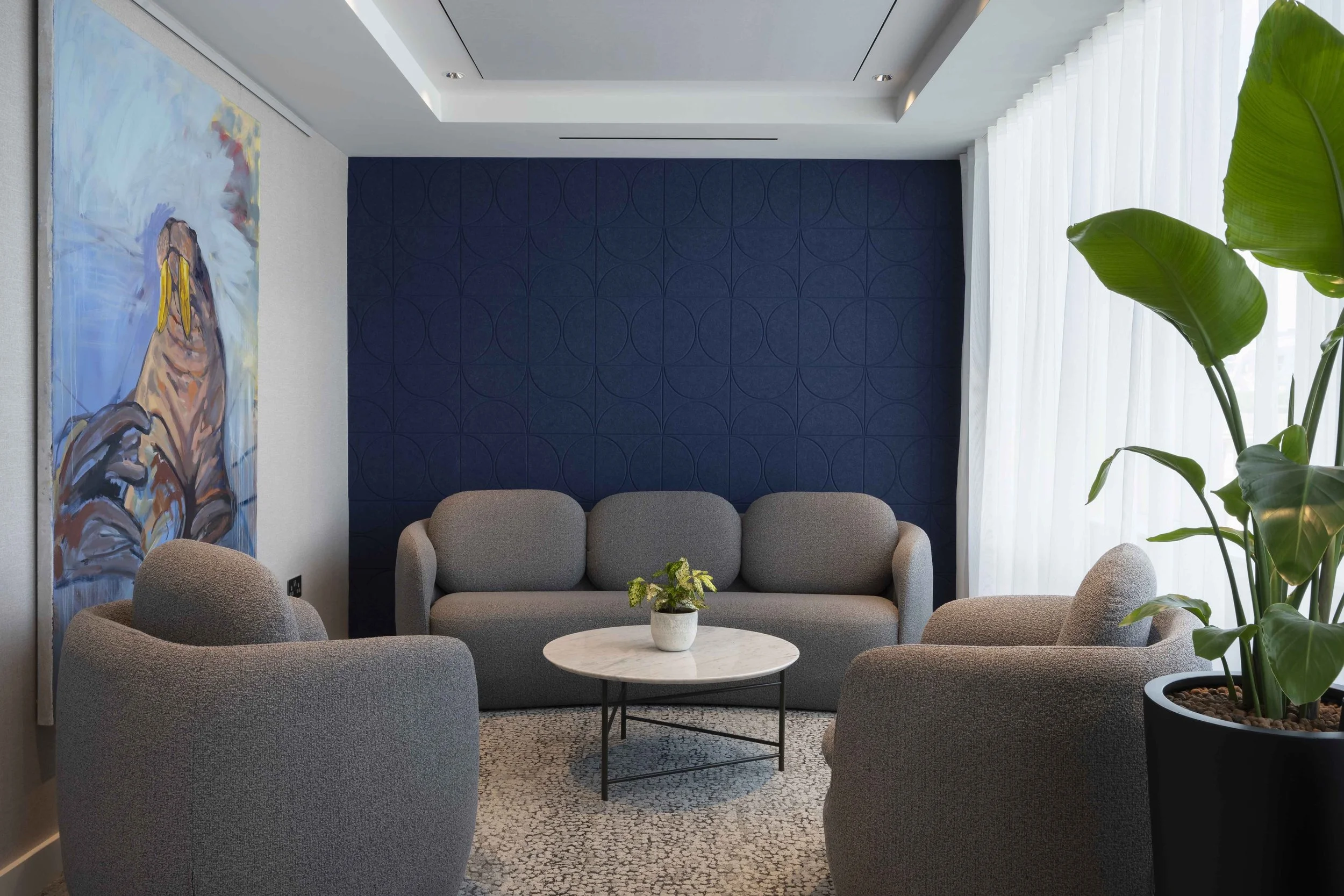 A modern living room with beige armchairs around a round marble coffee table, a blue accent wall, a large abstract painting, and a large potted plant near sheer white curtains.