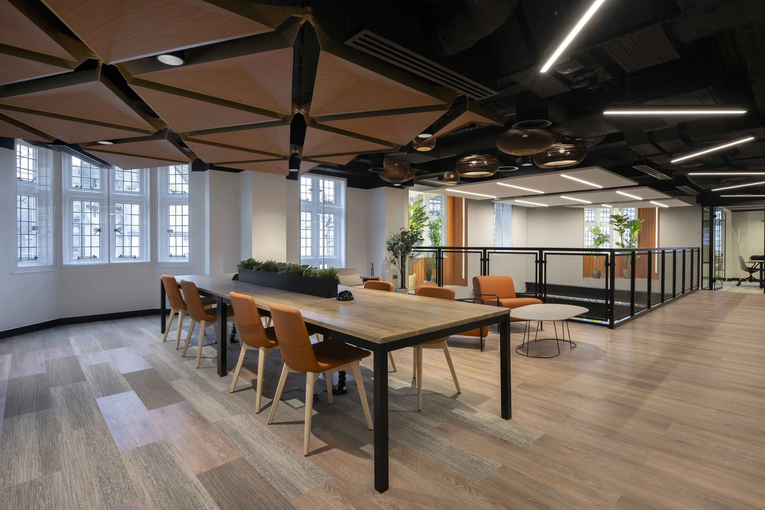 Modern office space with large windows, wooden ceiling panels, and a mix of seating areas including a long table with orange chairs, green plants, and unique ceiling fixtures.