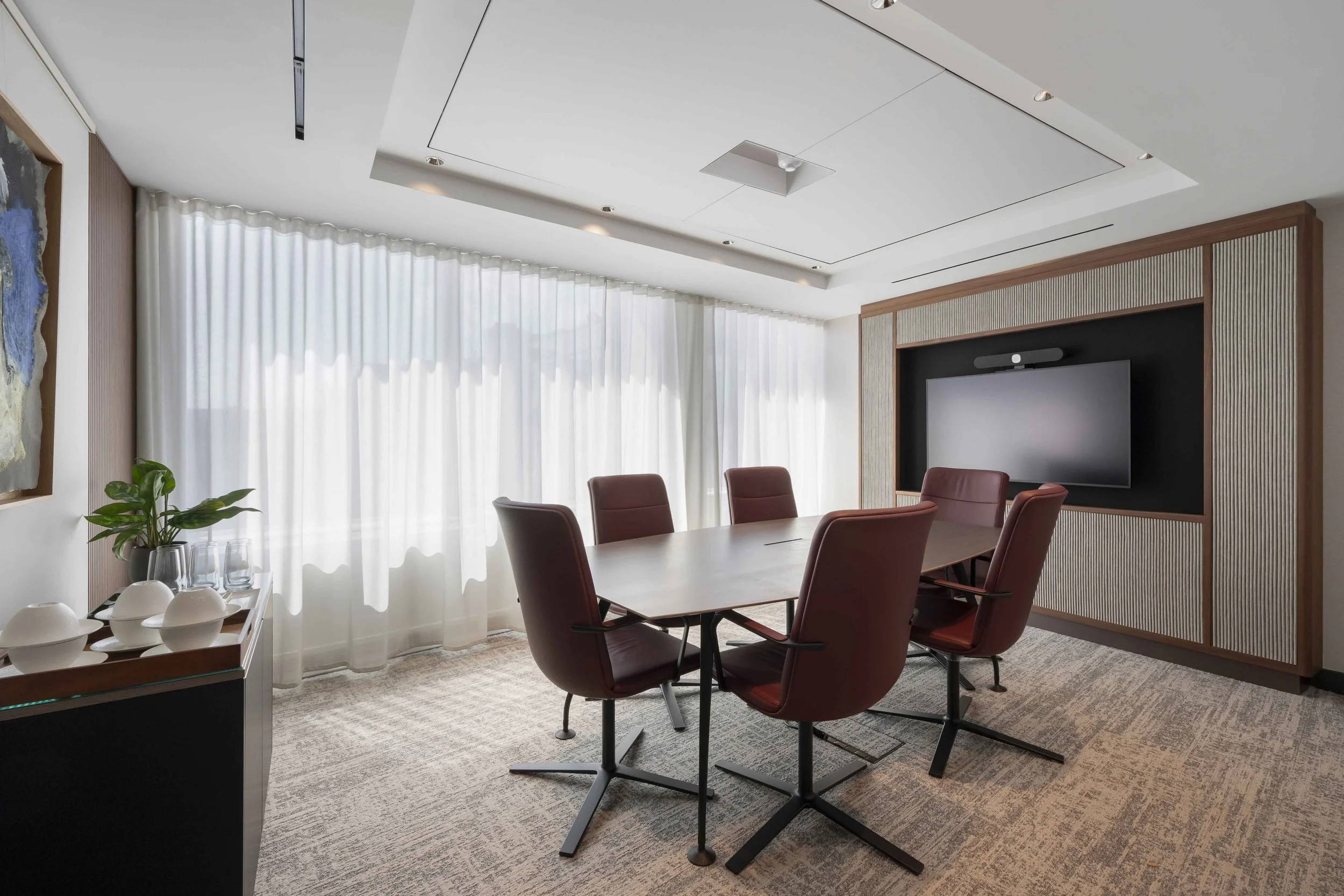 Modern conference room with a large wooden table surrounded by six maroon chairs. There is a large flat-screen TV on the wall, framed with wood paneling. White sheer curtains cover large windows, and a decorative painting is on the left wall. A side table with white bowls and a potted plant is in the foreground. The ceiling is white with embedded ceiling lights.