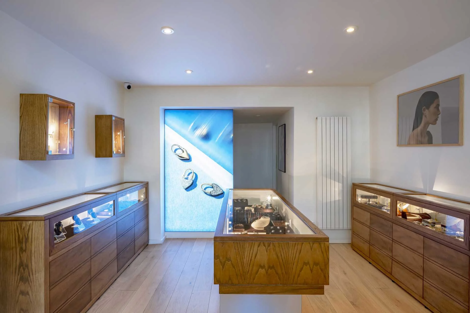 Interior of a jewelry store with display cases filled with jewelry on wooden counters, framed artwork on the walls, and a large handbag advertisement on the back wall.