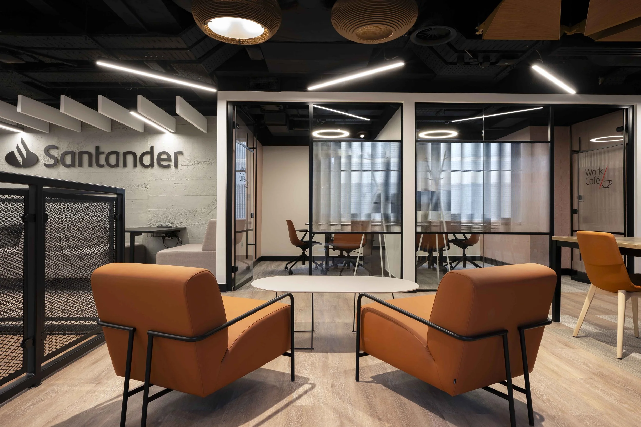 Modern office lounge area with orange armchairs, small round tables, glass conference room with frosted glass panels, and the Santander logo on a white brick wall.