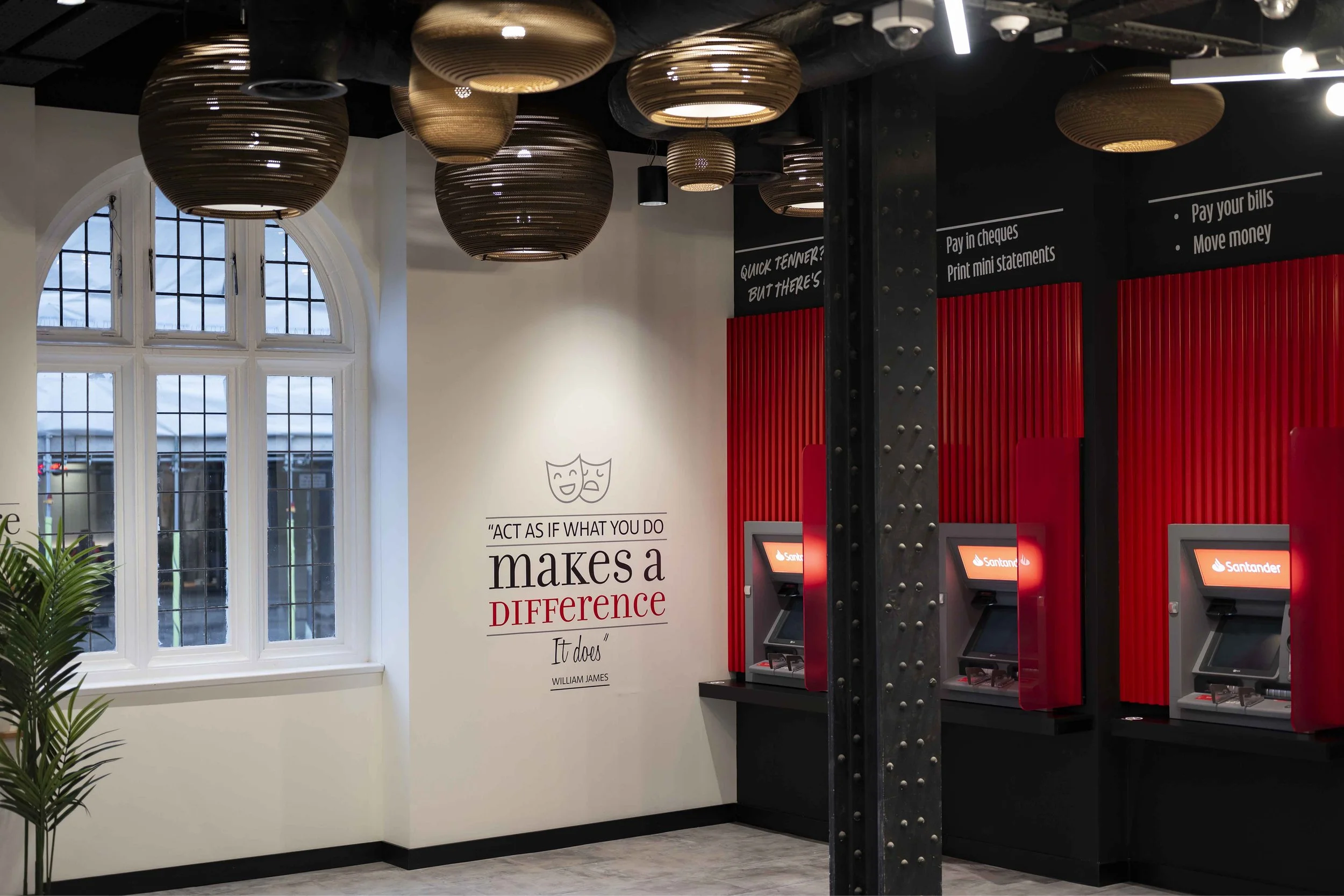 Interior view of a bank or financial institution with red ATMs, decorative ceiling lights, large windows, and a wall with quote art.