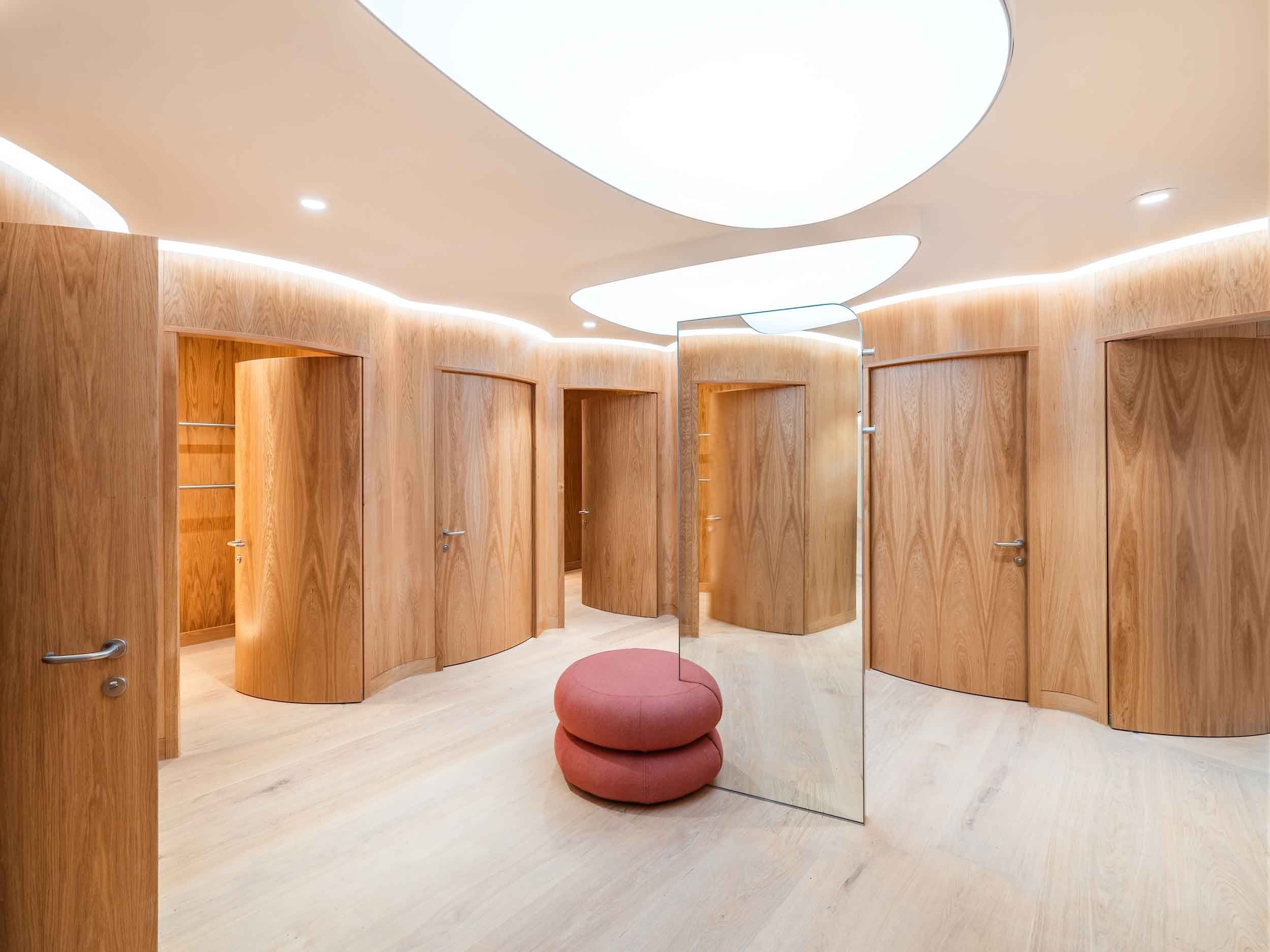 Fitting rooms with wooden doors and a round red stool in a clothing store with a modern ceiling light.