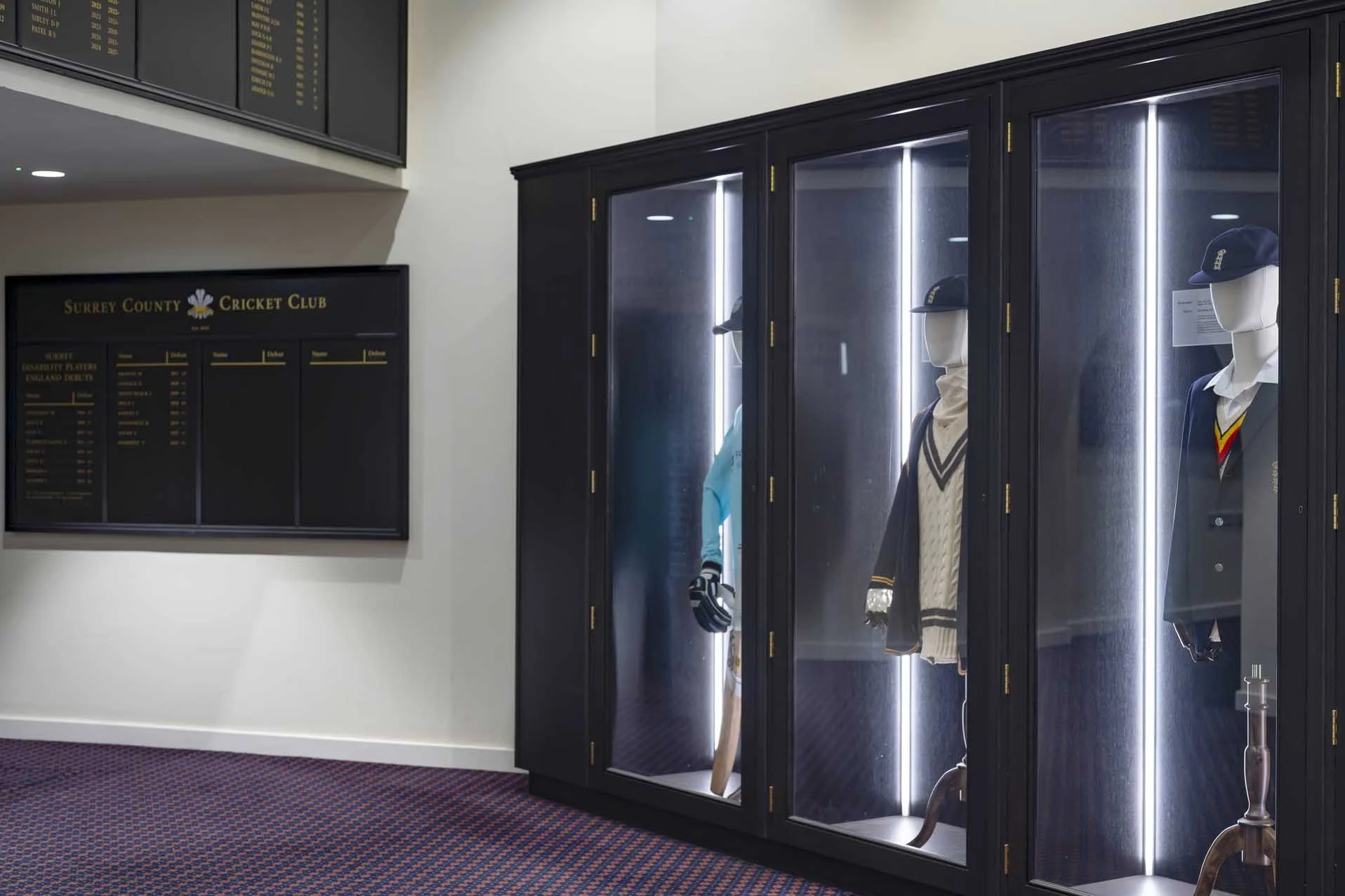 Glass display case with mannequins dressed in cricket uniforms, caps, and gloves inside. A mural or photo on the wall shows a cricket player. A black sign on the wall reads "Surrey County Cricket Club" with a list of players and stats.