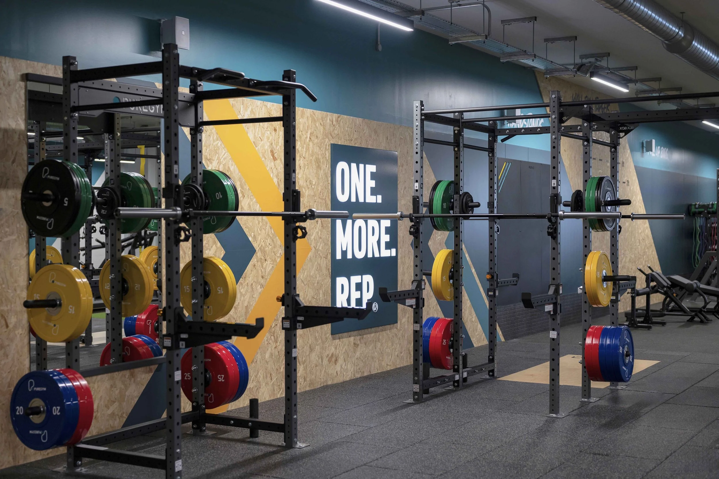 A gym with weightlifting equipment, including barbells and weight plates of various colors and sizes. The wall features motivational signage and modern industrial lighting.