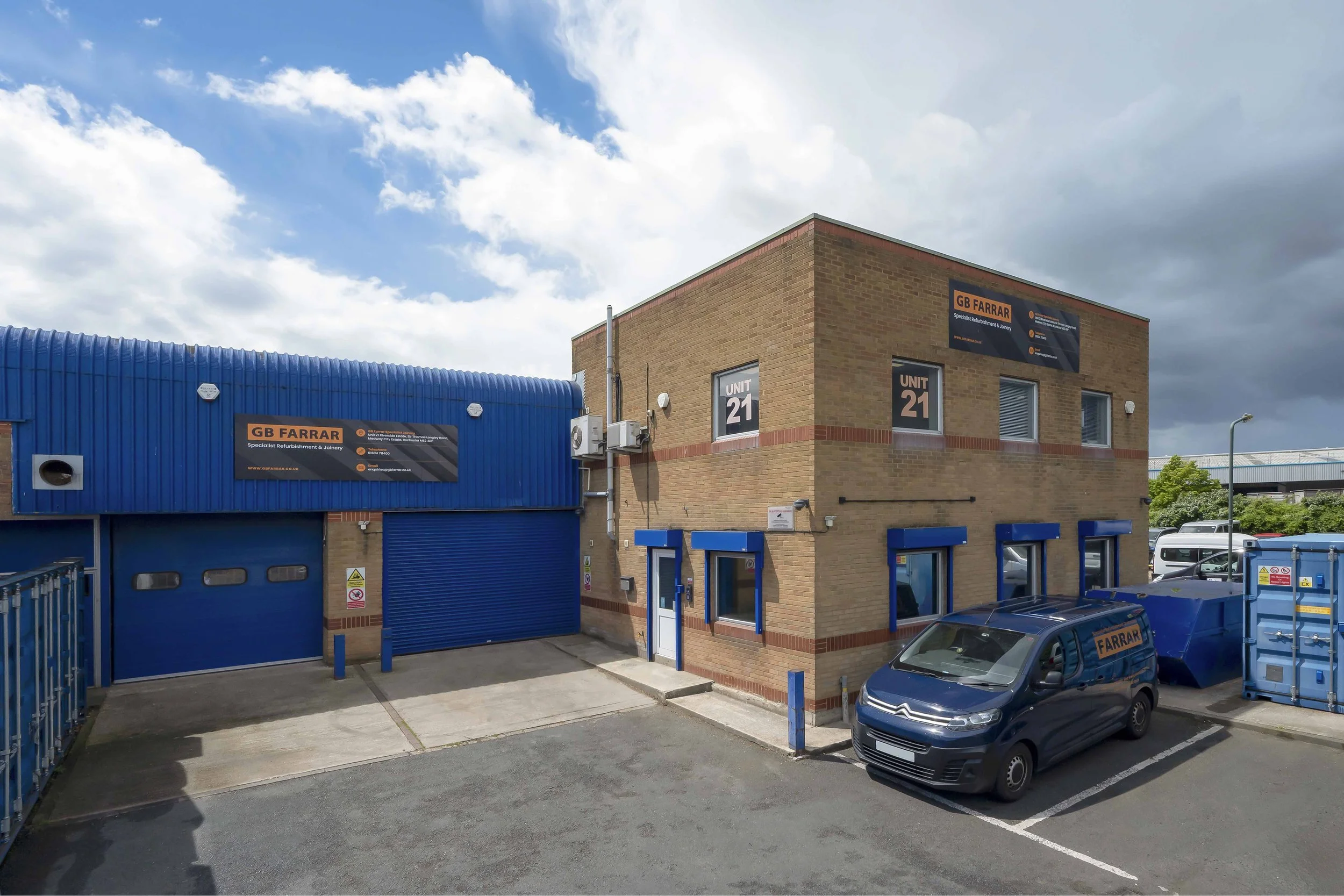 The image shows a commercial building with a brick exterior and signs that read 'UNIT 21' and 'GB FARRAR Specialized Refurbishment & Joinery.' There is a blue metal garage door, a parking lot with several cars, and a blue sky with scattered clouds overhead.