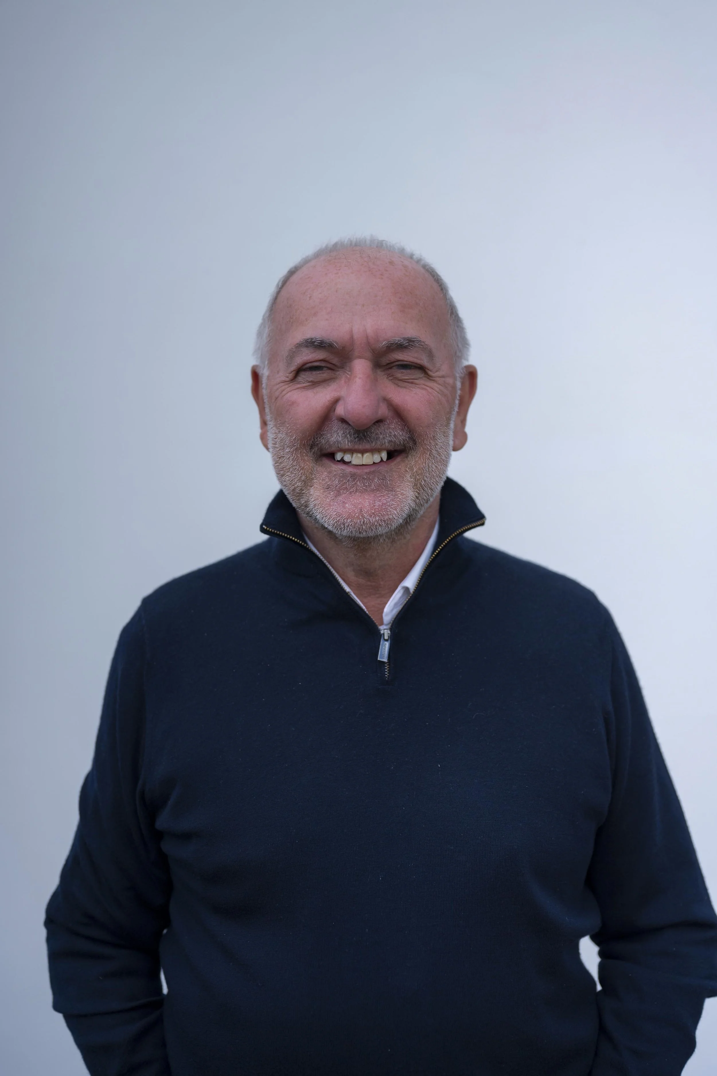 A smiling man with a beard and short gray hair wearing a dark zip-up sweater, standing against a plain light blue background.