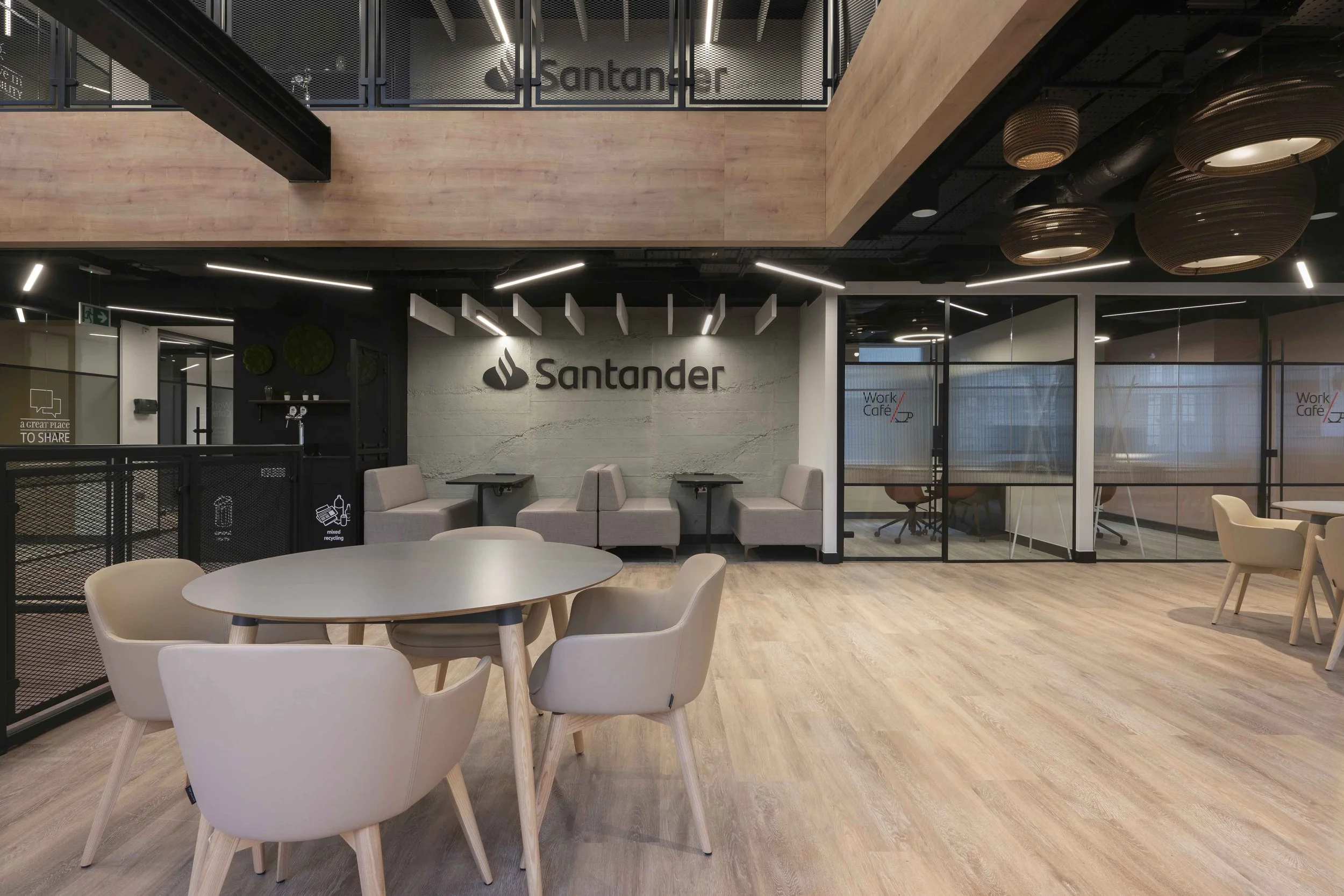 Modern office space with a seating area, round white table, beige chairs, glass-walled conference room, and wall-mounted Santander logo. Some decorative hanging lights and wood accents are visible.