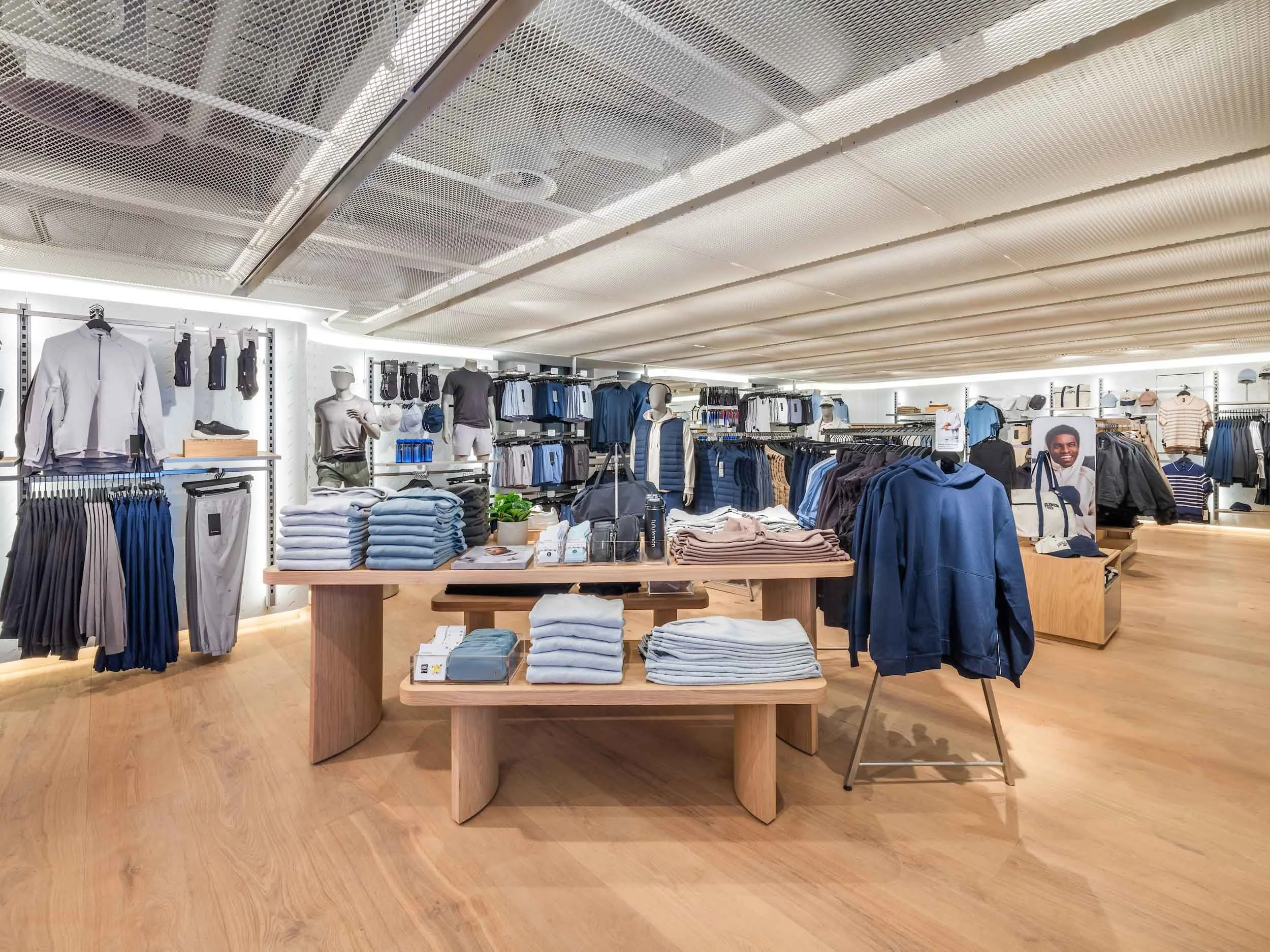Inside a retail clothing store displaying men's apparel including shirts, jackets, shorts, and accessories on mannequins and racks, with a wooden display table in the foreground.