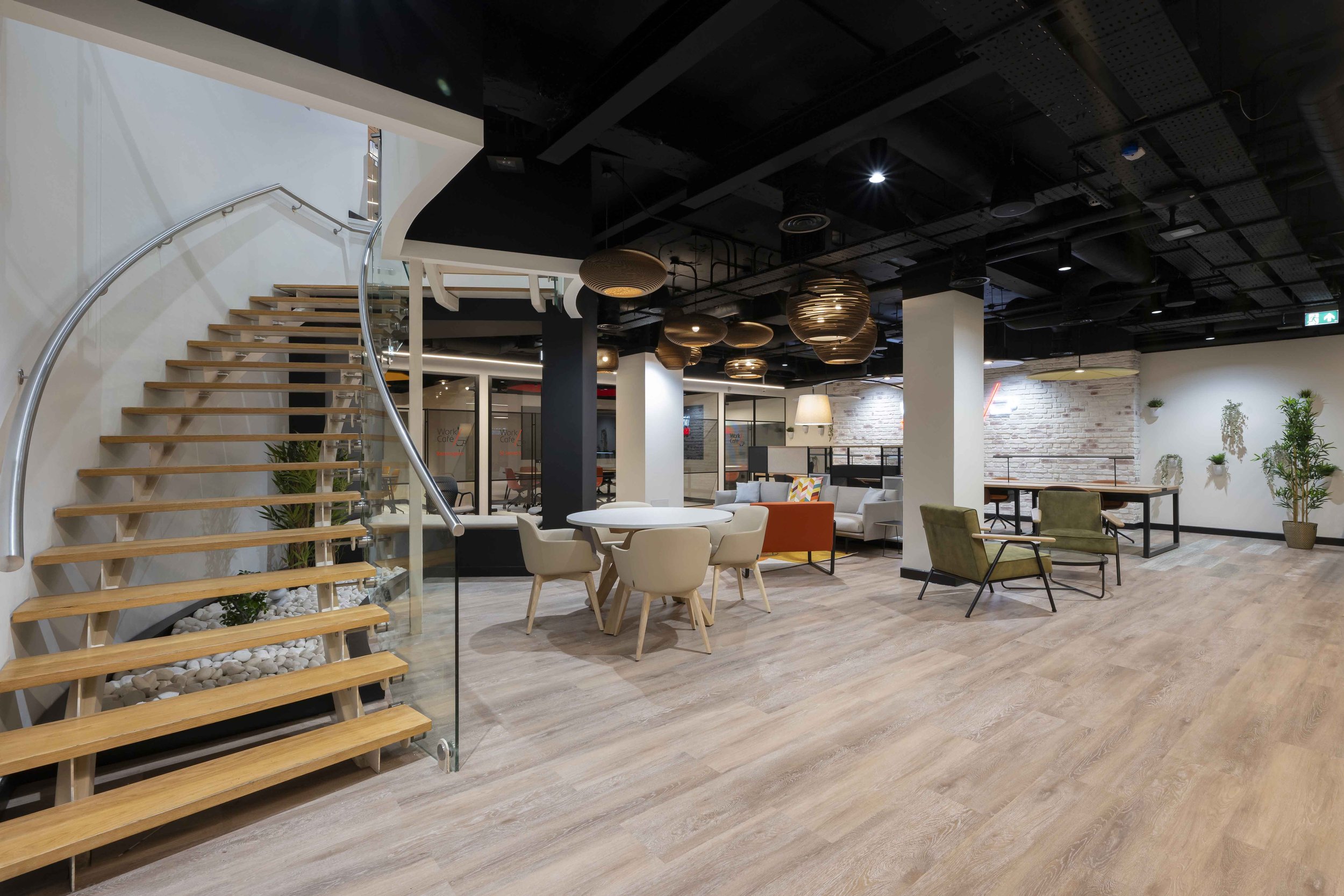 Modern office lobby with sitting area, wooden staircase with glass railing, hanging pendant lights, white brick wall, various chairs and plants, and a black ceiling with exposed ductwork.