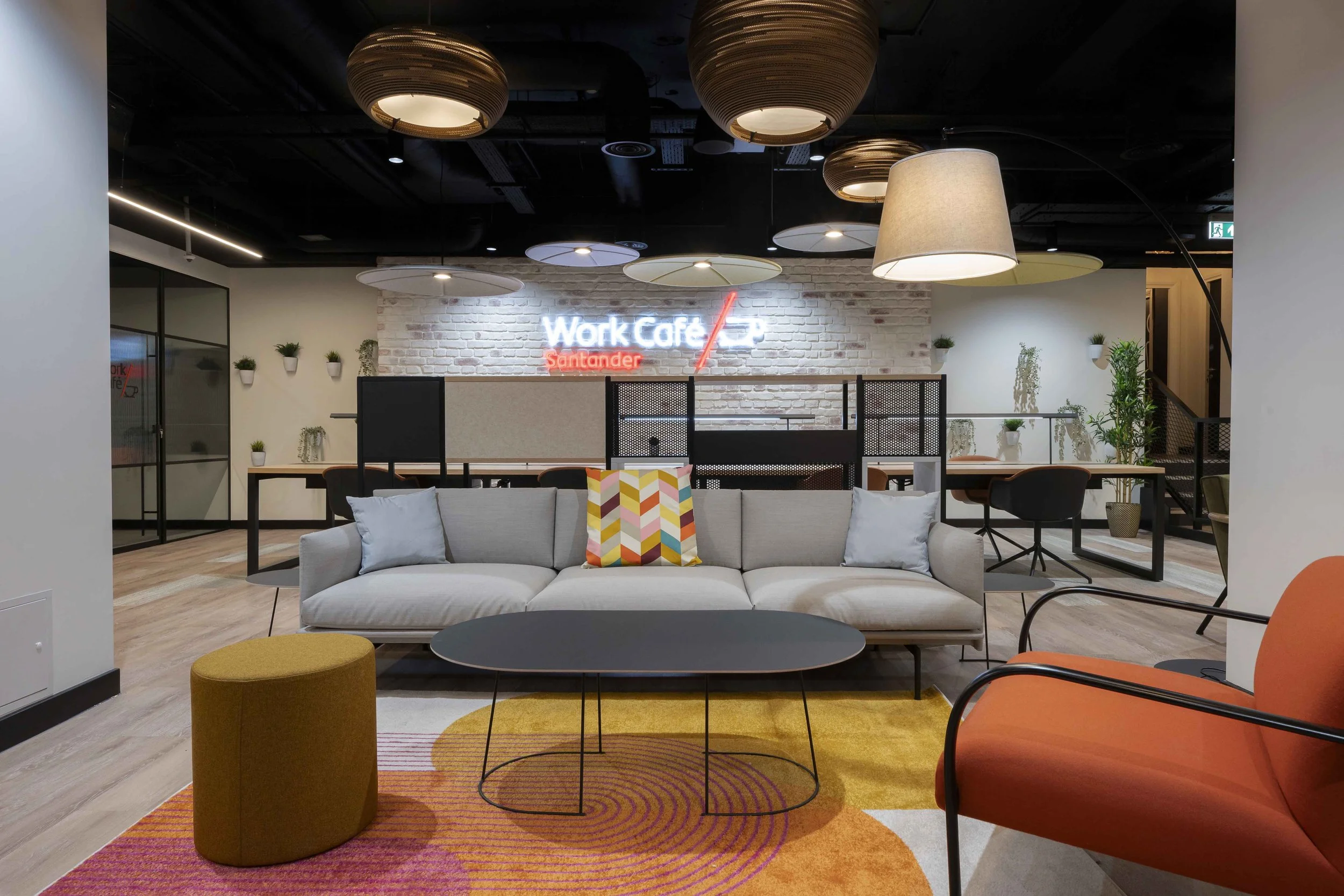 A modern office lounge area with a white sofa adorned with colorful cushions, a yellow ottoman, a gray round coffee table, and a red chair. The background features a brick accent wall with a neon sign reading 'Work Café/Santander'. The ceiling has ro