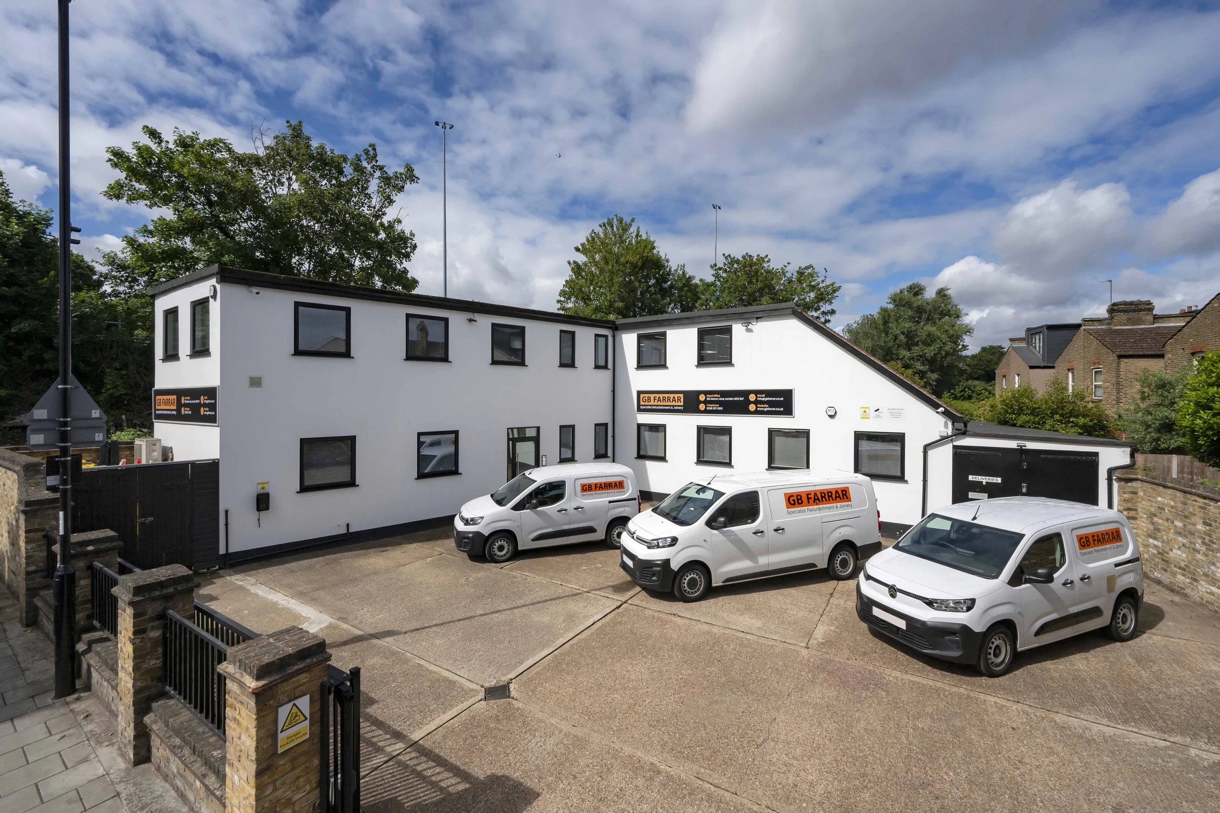 A white commercial building with several windows and signs for GB Farrar, a specialist refurbishment and joinery company, parked with multiple white delivery vans in front. The sky above is partly cloudy with blue patches, and there are trees and neighboring houses in the background.