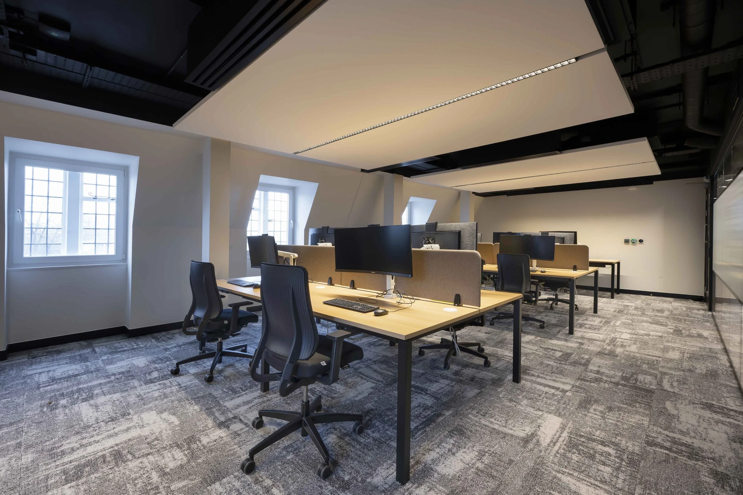 Modern office space with large windows, multiple desks with chairs and computer monitors, overhead lighting, and a gray patterned carpet.